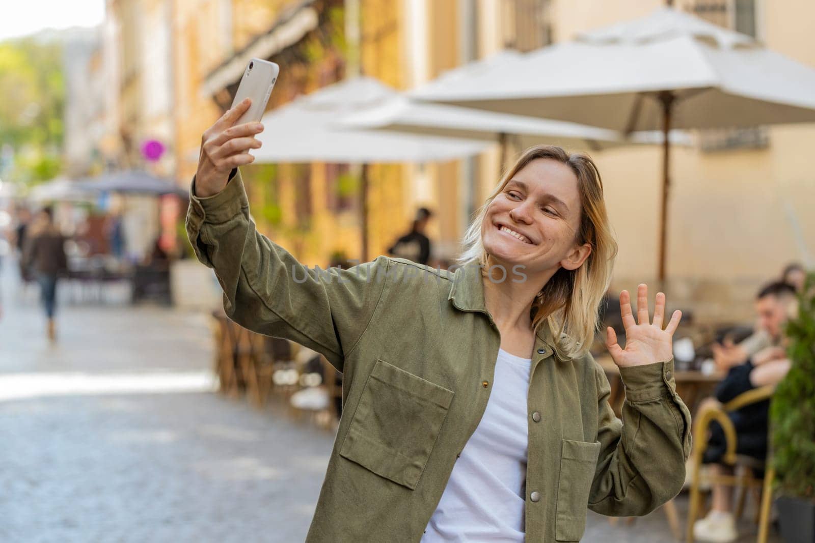 Young woman blogger taking selfie on smartphone, communicating video call online with subscribers, recording stories for social media vlog outdoors. Tourist traveler girl walking on urban city street.
