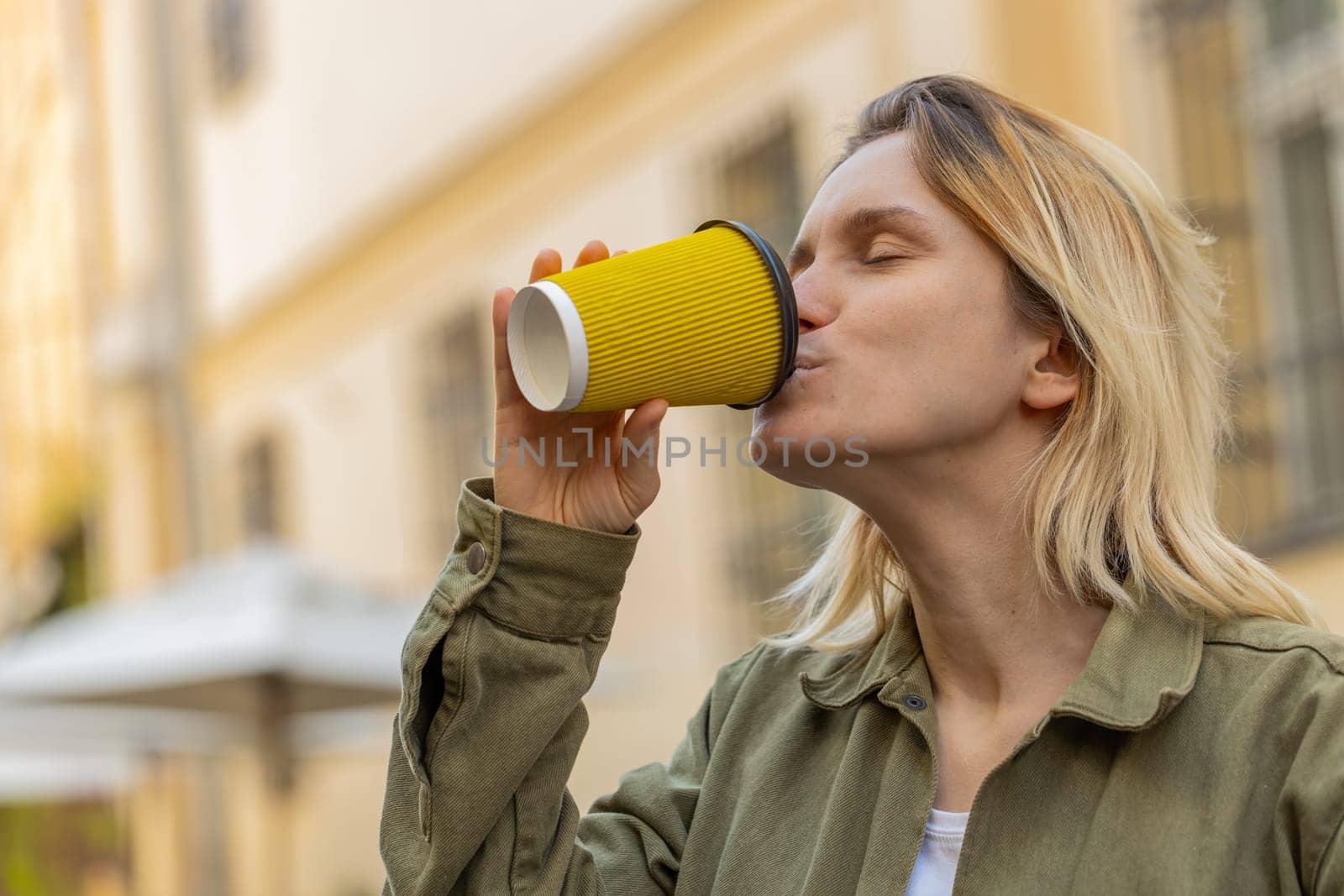 Happy Caucasian young woman enjoying morning coffee hot drink and smiling outdoors. Relaxing, taking a break. Girl walking through urban city center street, drinking coffee to go. Town lifestyles.