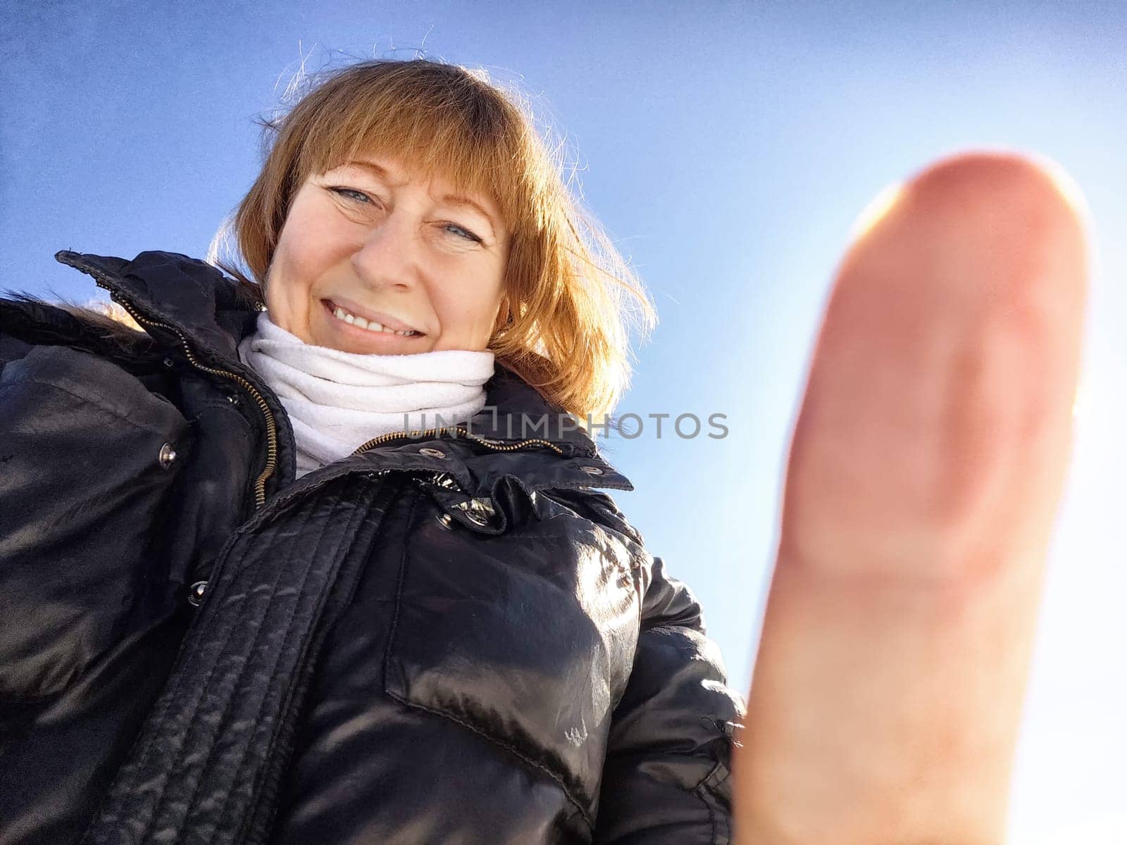 A cheerful middle aged woman in a winter coat taking selfie on nature outdoors in sunny day with blue sky