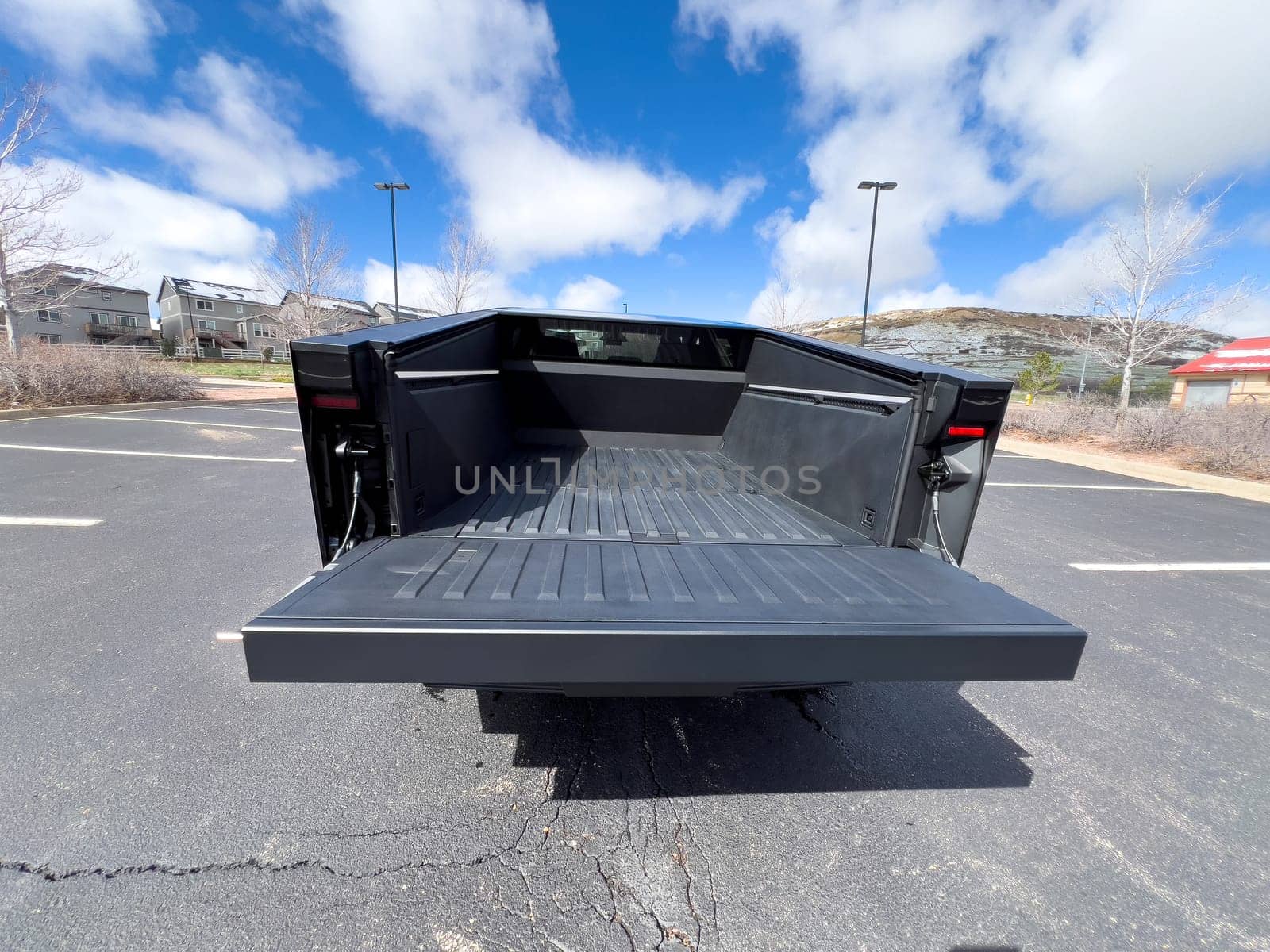Denver, Colorado, USA-March 28, 2024-The distinctive rear end of a Tesla Cybertruck, captured in an empty parking lot, stands out against a backdrop of suburban homes under a blue sky with fluffy clouds.