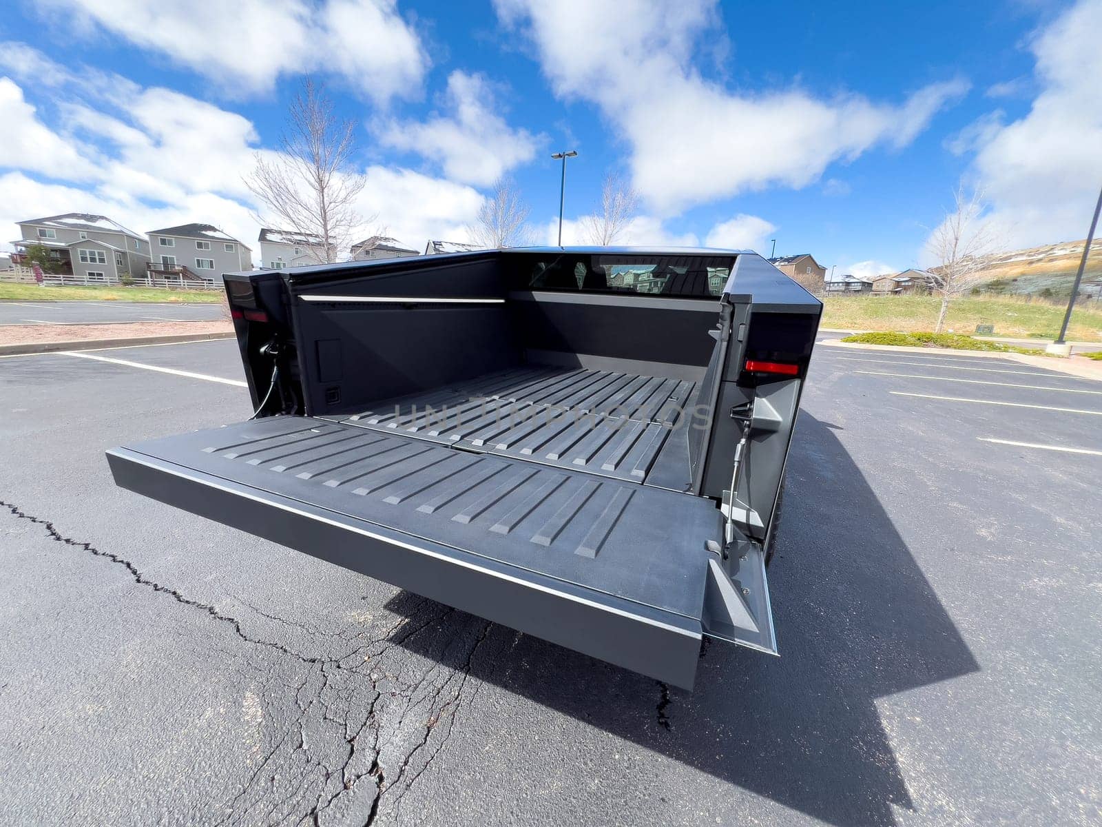 Denver, Colorado, USA-March 28, 2024-The distinctive rear end of a Tesla Cybertruck, captured in an empty parking lot, stands out against a backdrop of suburban homes under a blue sky with fluffy clouds.