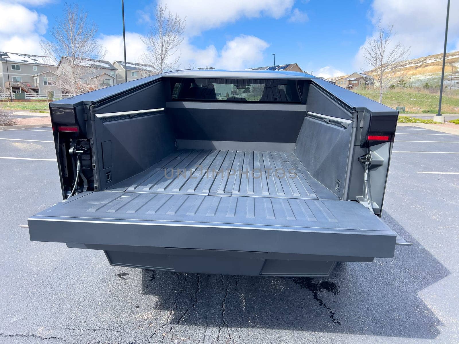 Denver, Colorado, USA-March 28, 2024-The distinctive rear end of a Tesla Cybertruck, captured in an empty parking lot, stands out against a backdrop of suburban homes under a blue sky with fluffy clouds.