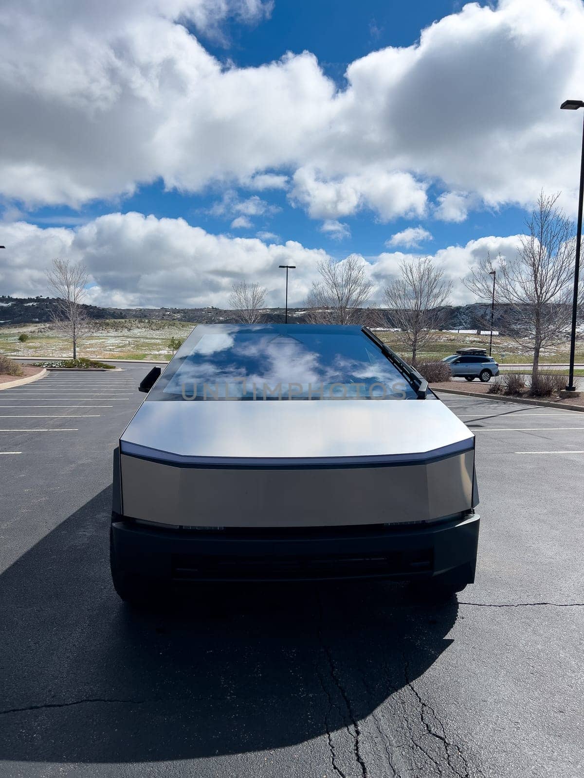 Denver, Colorado, USA-March 28, 2024-The front profile of a Tesla Cybertruck is displayed, showcasing its futuristic lines and robust design, parked in an open-air parking lot with a dynamic sky overhead.