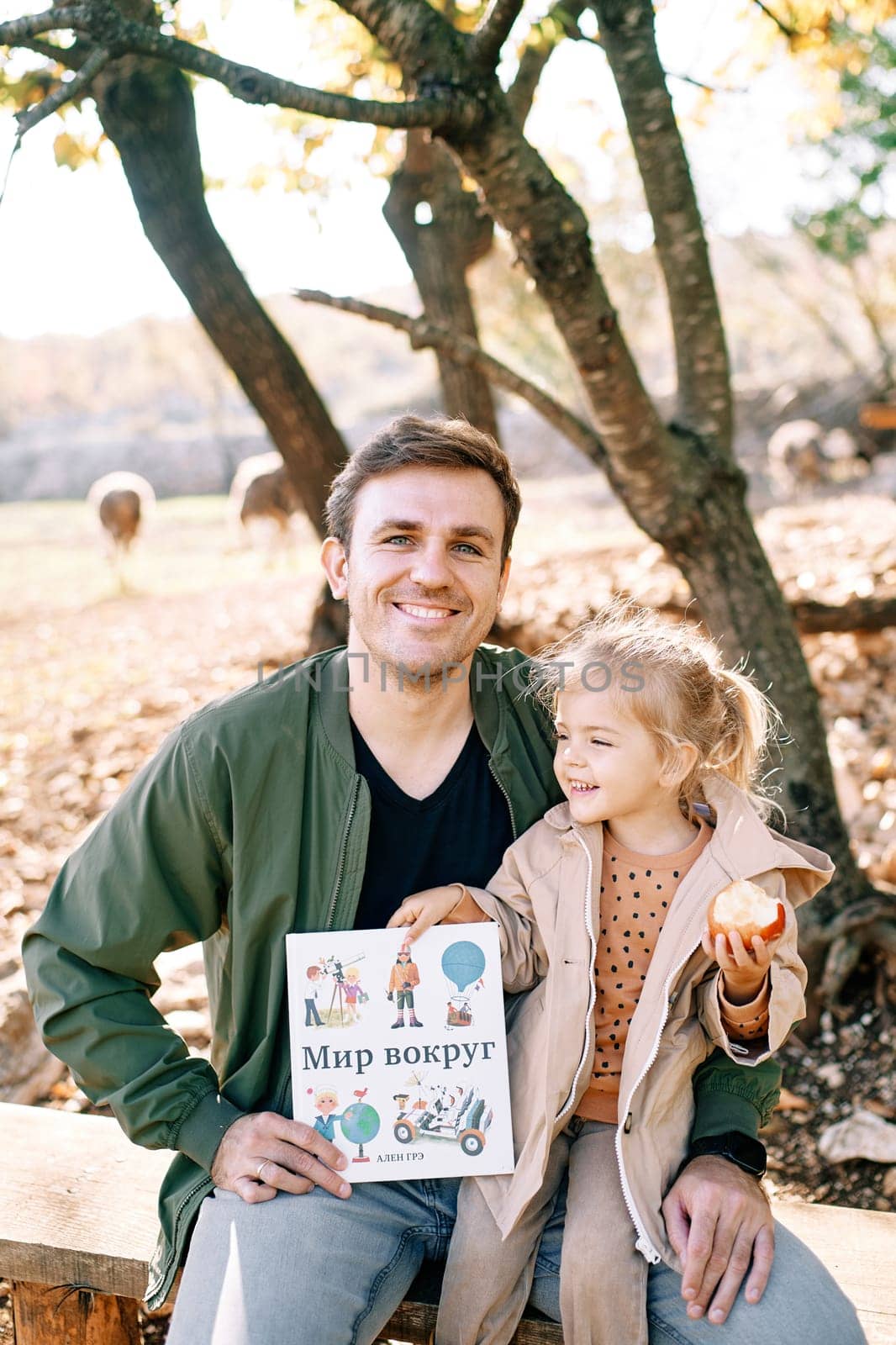 Smiling dad with a little girl on his lap sits on a bench with a book in his hands. Caption: World around. High quality photo