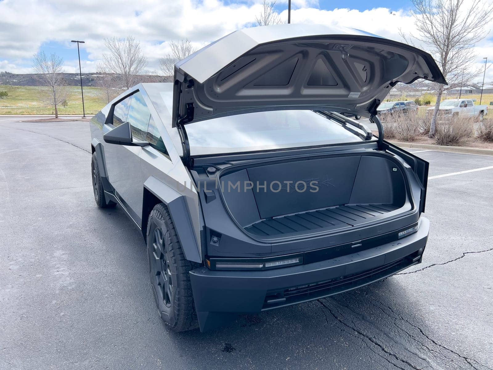 Denver, Colorado, USA-March 28, 2024-The front profile of a Tesla Cybertruck is displayed, showcasing its futuristic lines and robust design, parked in an open-air parking lot with a dynamic sky overhead.