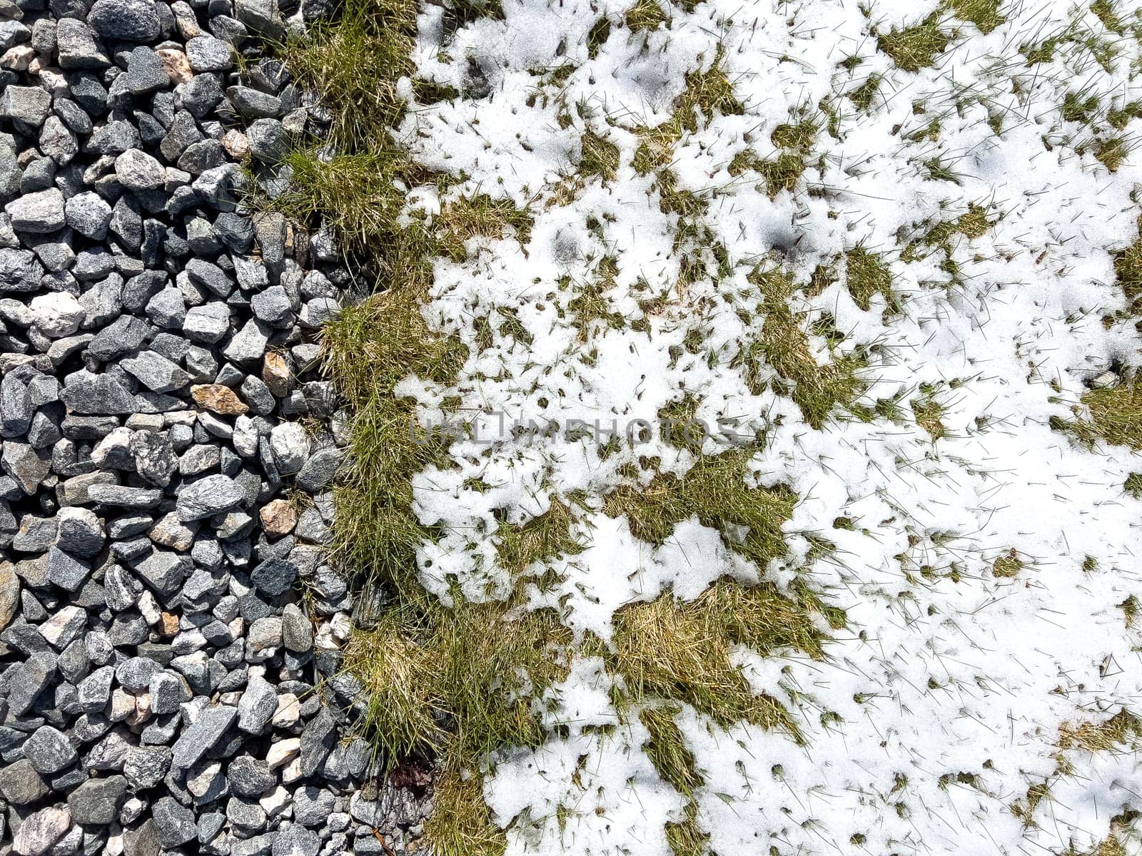 An image showing the remnants of a spring snowstorm on a suburban landscape, where melting snow meets the contrasting textures of gravel and green grass.