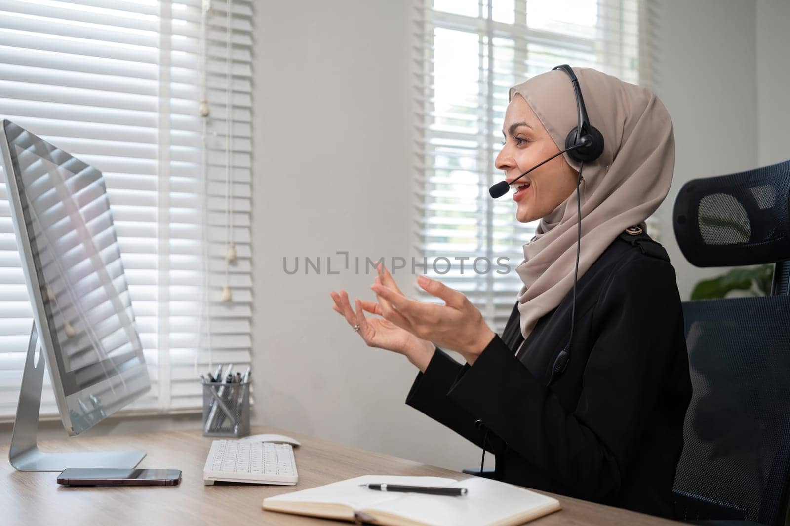 Call center worker, young Muslim woman wearing hijab, talking to customer on call phone on computer in customer service office.