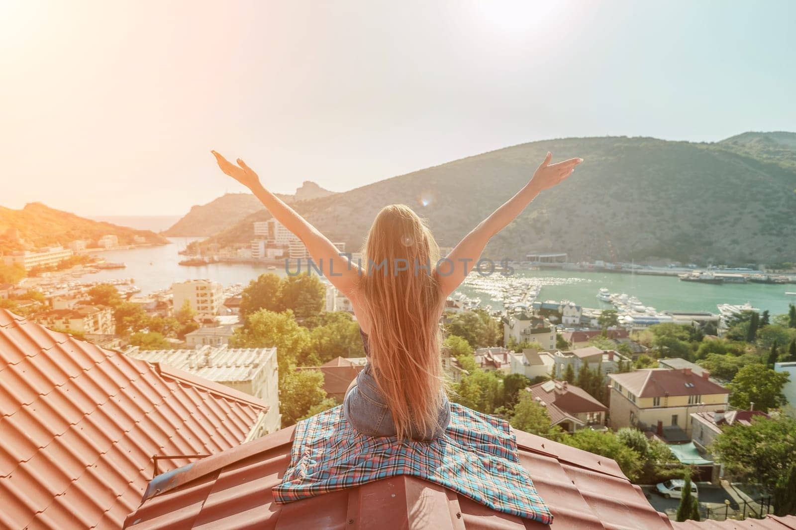 Woman sits on rooftop with outstretched arms, enjoys town view and sea mountains. Peaceful rooftop relaxation. Below her, there is a town with several boats visible in the water by Matiunina
