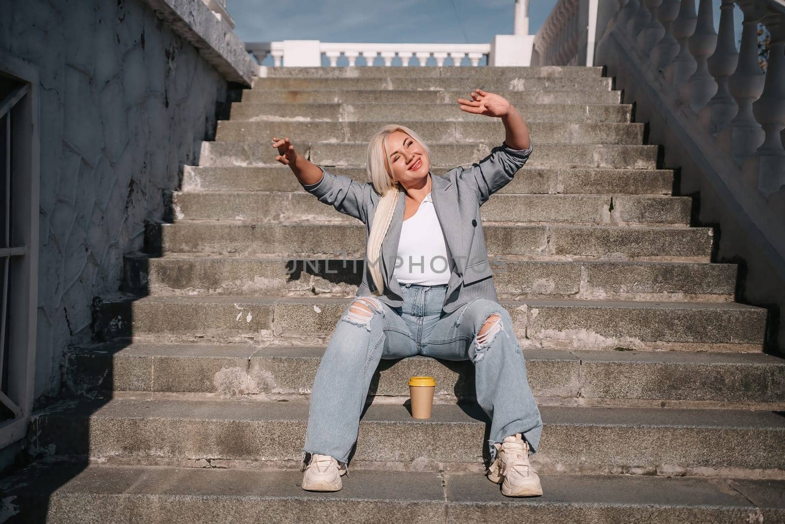 A woman in jeans and a jacket sits on a set of stairs. She is smiling and holding a cup