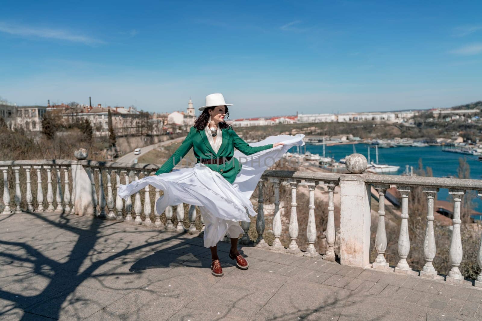Woman walks around the city, lifestyle. Happy woman in a green jacket, white skirt and hat is sitting on a white fence with balusters overlooking the sea bay and the city. by Matiunina