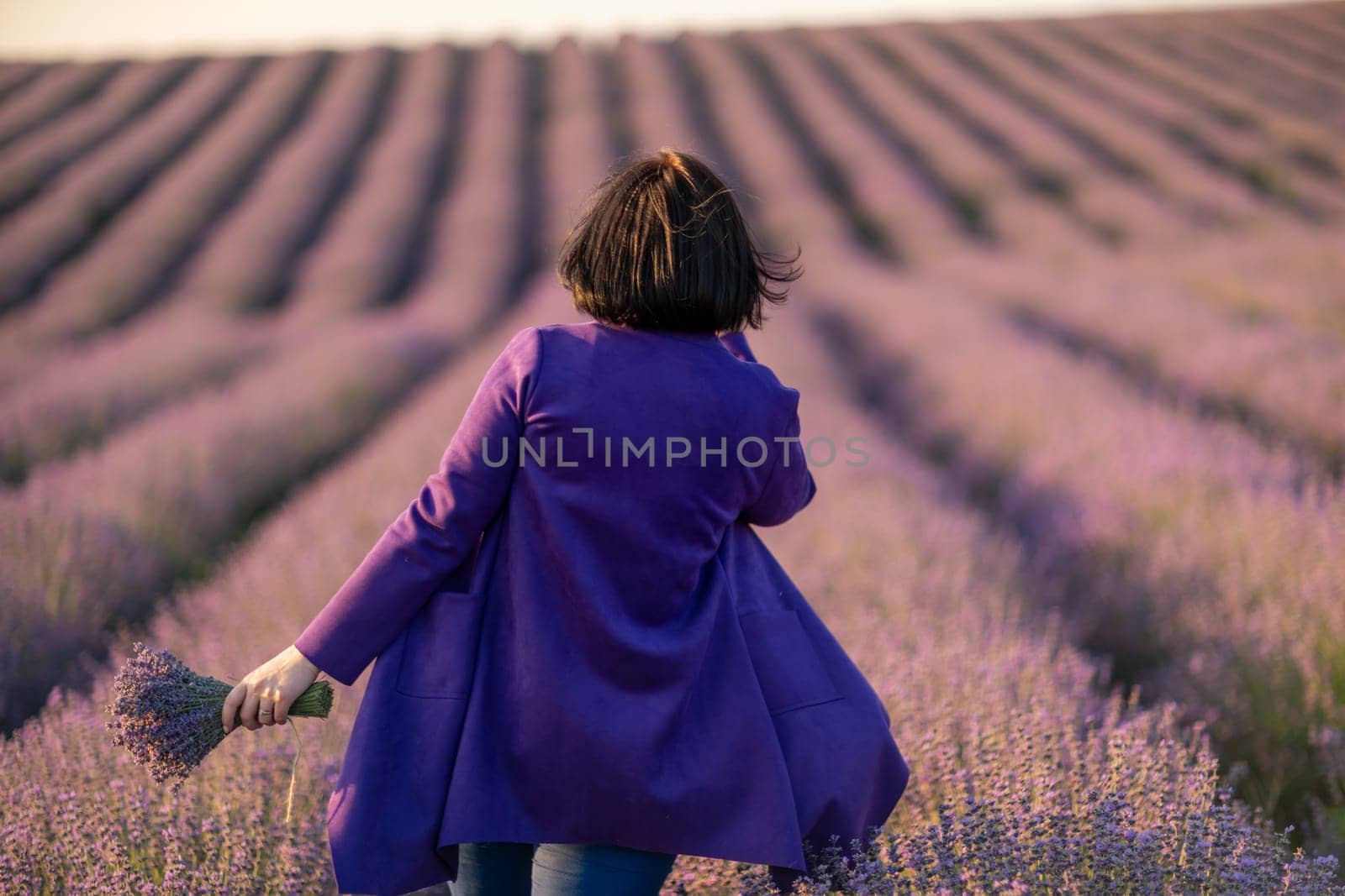 A woman in a purple coat is walking through a field of lavender. The scene is serene and peaceful, with the purple flowers creating a calming atmosphere. The woman is enjoying her time in the field. by Matiunina