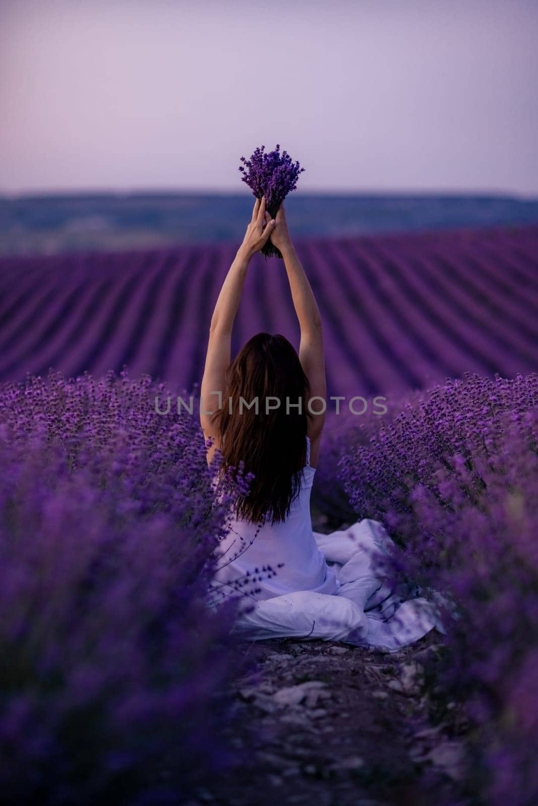 A woman is sitting in a field of lavender flowers. She is holding a bouquet of flowers in her hands. The scene is serene and peaceful, with the purple flowers creating a calming atmosphere. by Matiunina