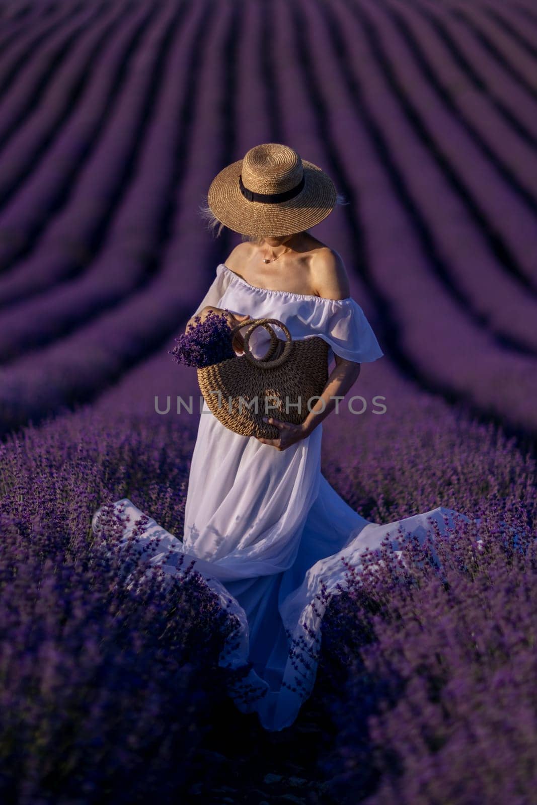 A woman is standing in a field of purple lavender flowers, wearing a straw hat and holding a basket of flowers