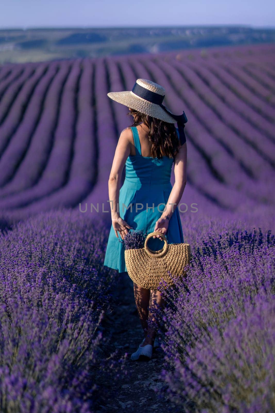 A woman wearing a straw hat and a blue dress is walking through a field of lavender. She is holding a basket and she is enjoying the scenery. by Matiunina