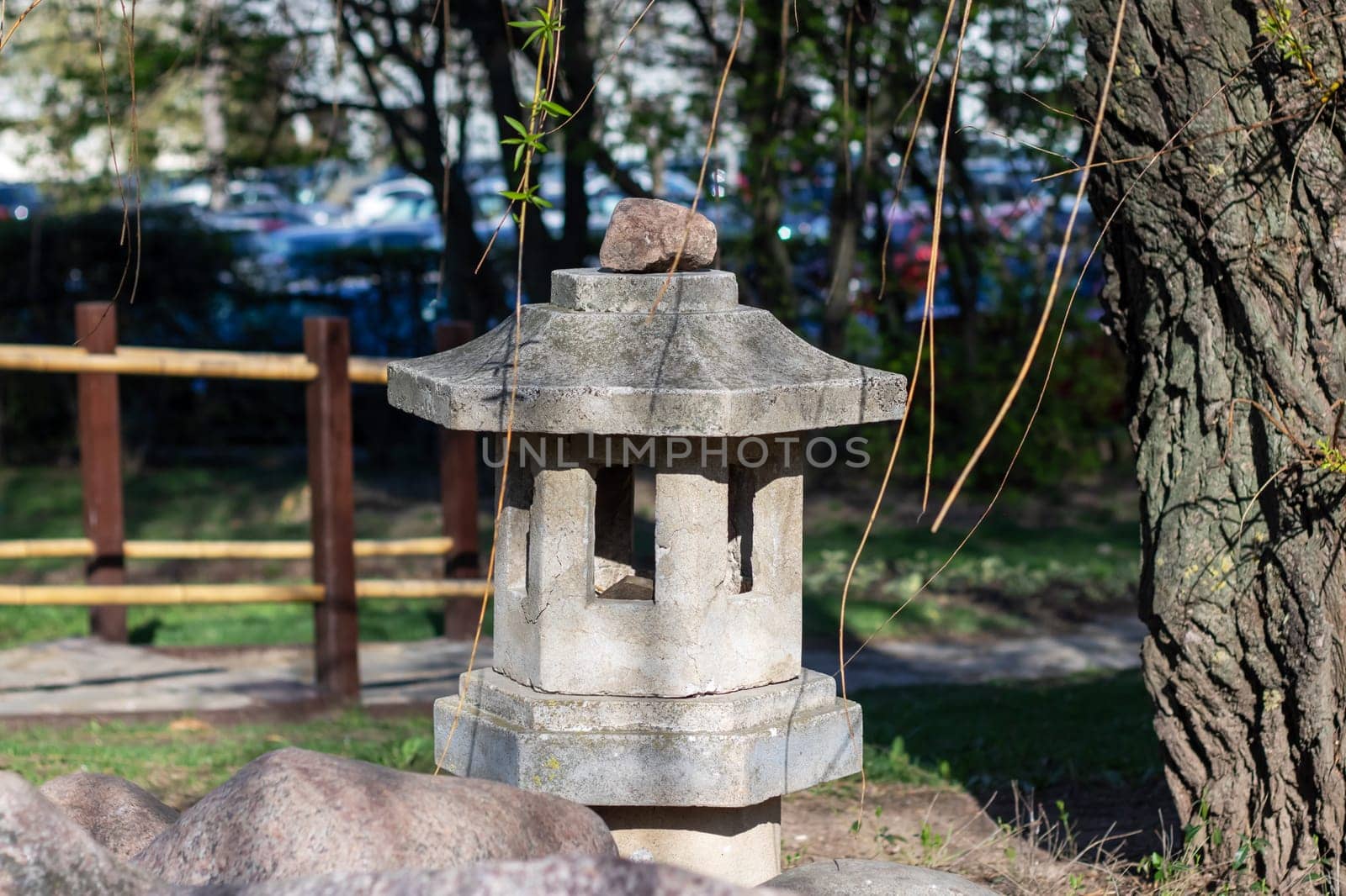 A stone lantern sits beside a tree in a park, adding a touch of art to the natural landscape. The lantern contrasts beautifully with the green grass and wood trunk of the tree