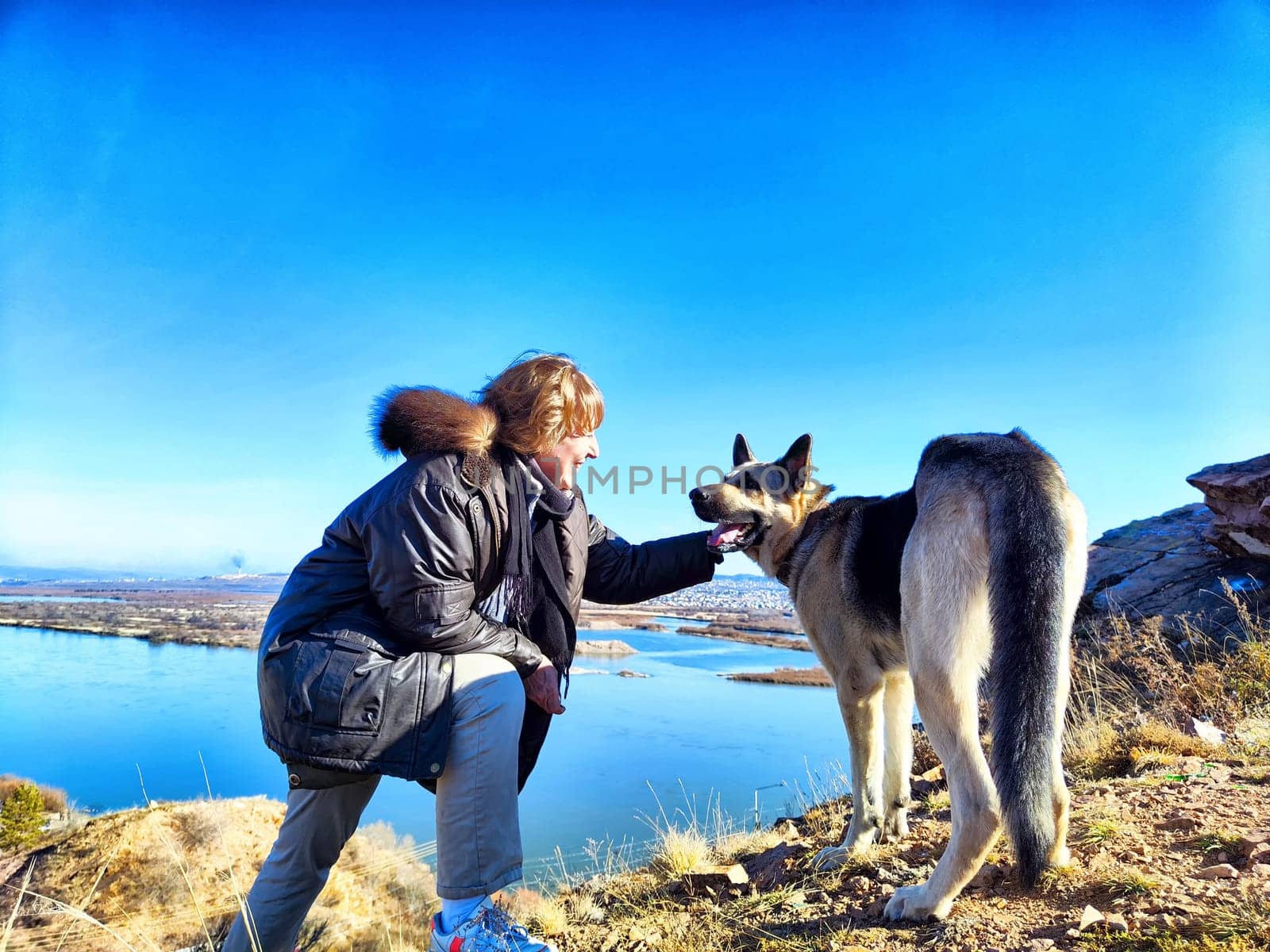 Adult girl with shepherd dog taking selfie near water of lake or river in mountain. Middle aged woman and big pet on nature. Friendship, love and fun