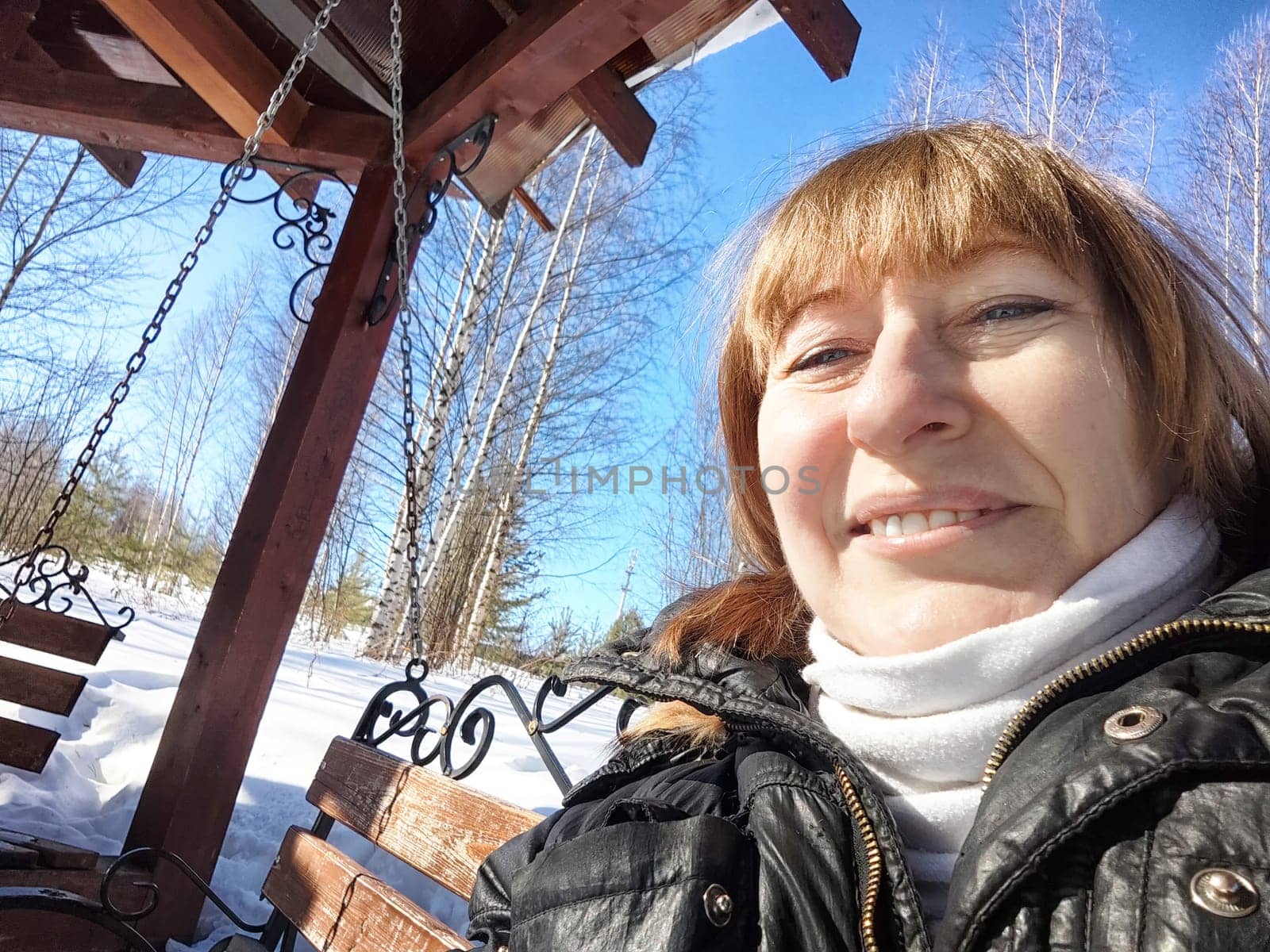 A cheerful middle aged woman in a winter coat taking selfie on the swing on nature outdoors in sunny day with blue sky