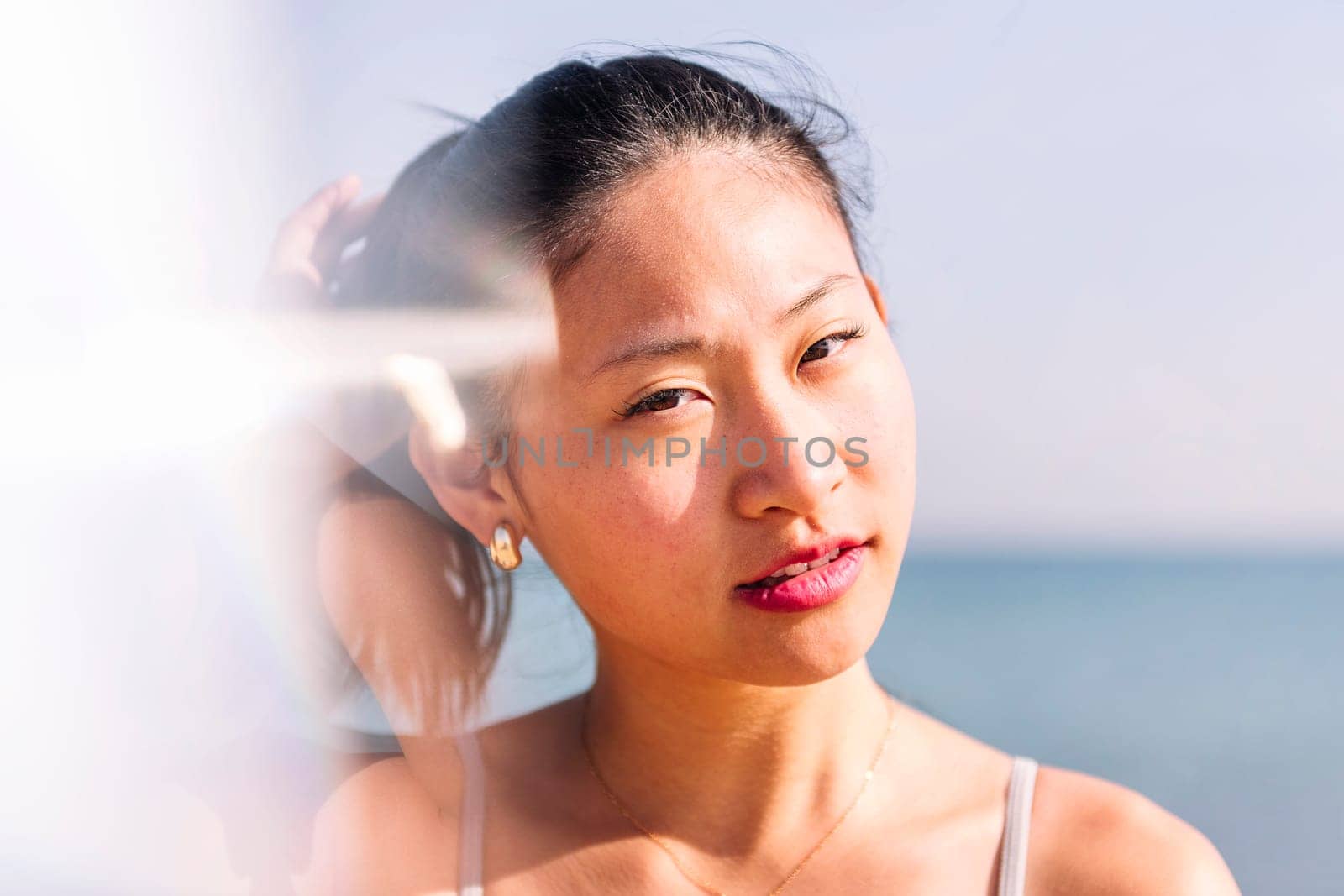 young contemplative asian woman at beach looking at camera, concept of beauty and purity, flare effect