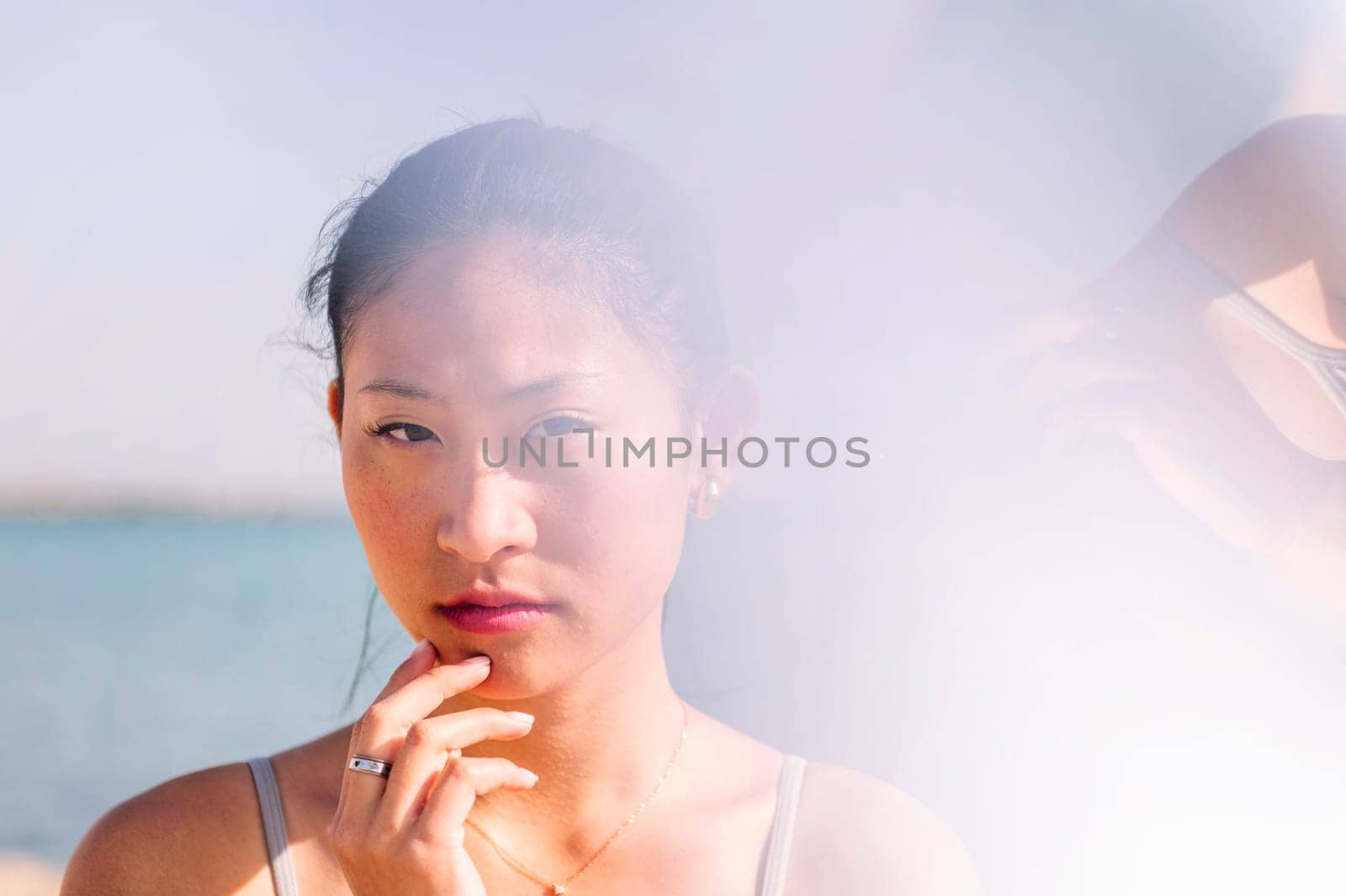 young contemplative asian woman touching face and looking at camera, concept of beauty and purity, flare effect