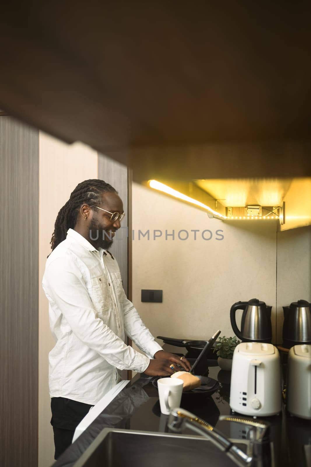 Happy African man in casual clothes using digital tablet on kitchen counter by prathanchorruangsak