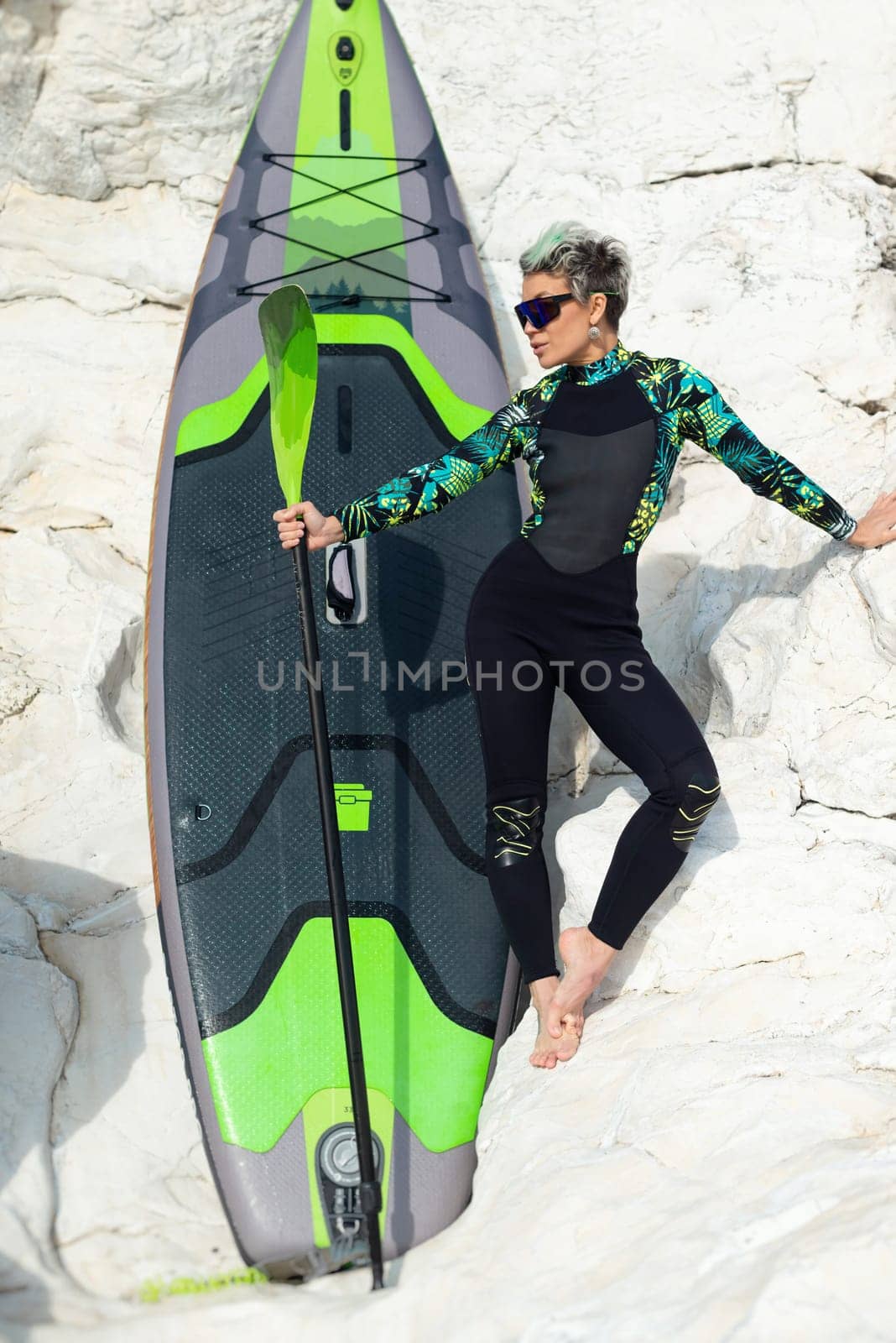 A beautiful sup surfer girl in a wetsuit on the ocean shore against a beautiful blue sky poses sexually with a paddle by Rotozey