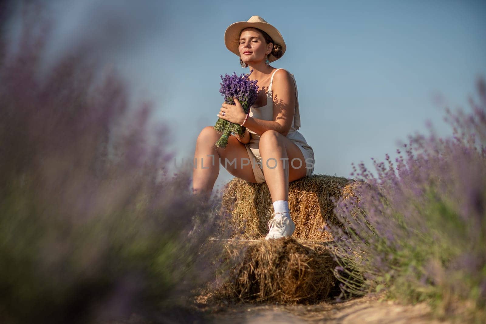 woman sitting in a field of lavender and wearing a straw hat. She is smiling and holding a bouquet of flowers. Scene is peaceful and serene, as the woman is surrounded by the beauty of nature by Matiunina