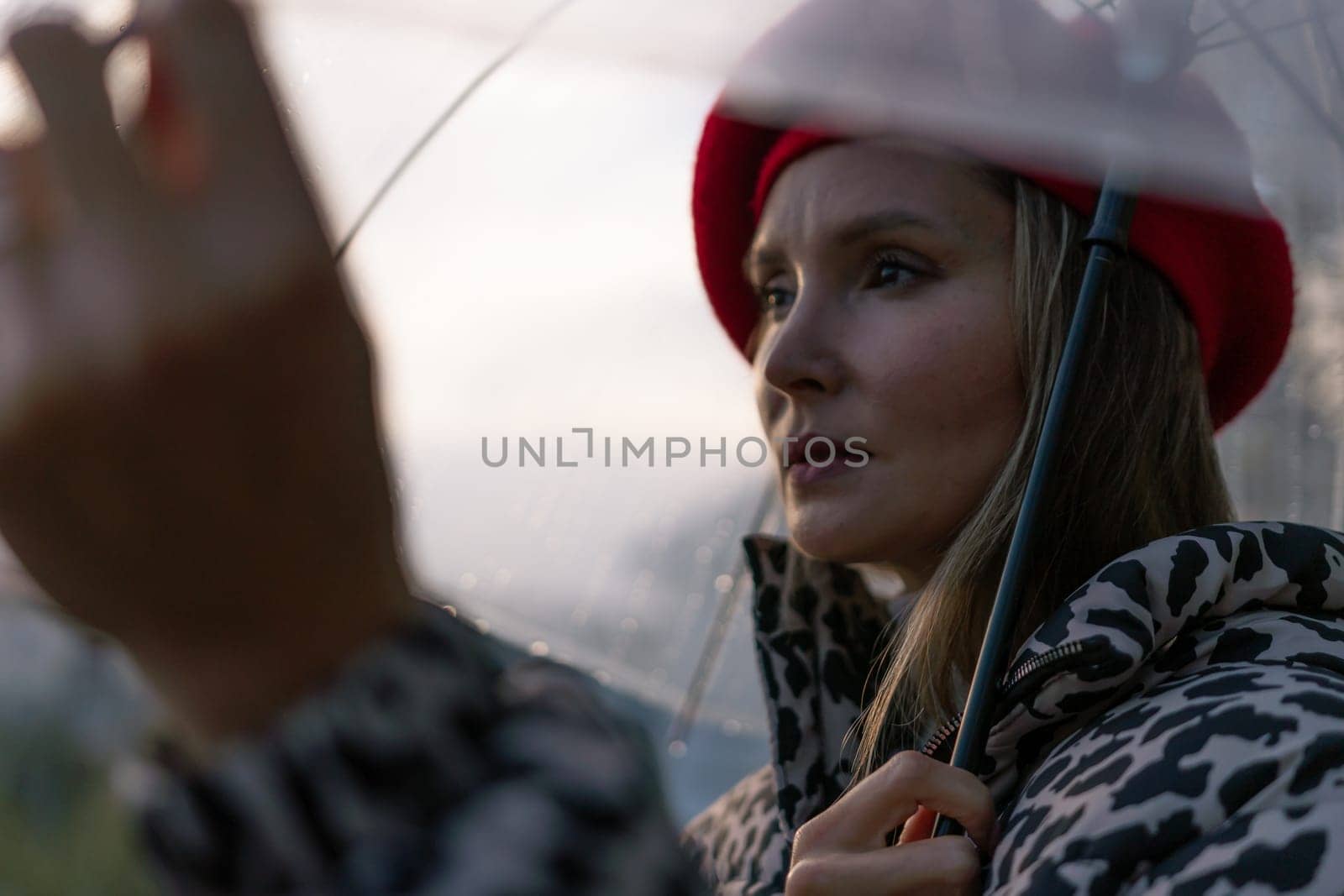 A woman wearing a red hat and a leopard print coat is holding a clear umbrella. The umbrella is open, and the woman is looking up at the sky. The scene has a calm and peaceful mood