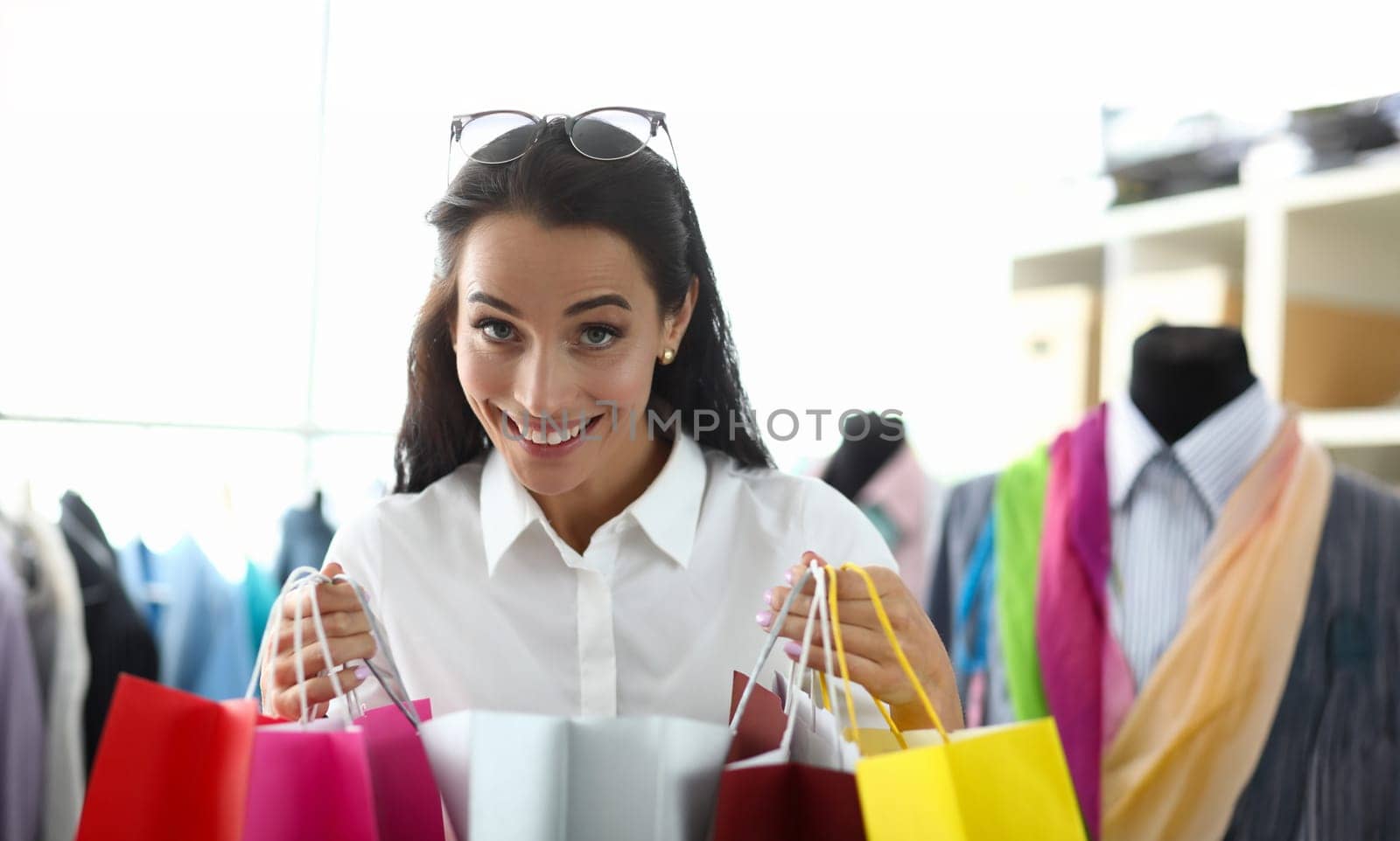 Portrait of smiling woman standing in modern clothes shop and holding many multicolored packets of new outfits and looking at camera with happiness. Shopping sale concept