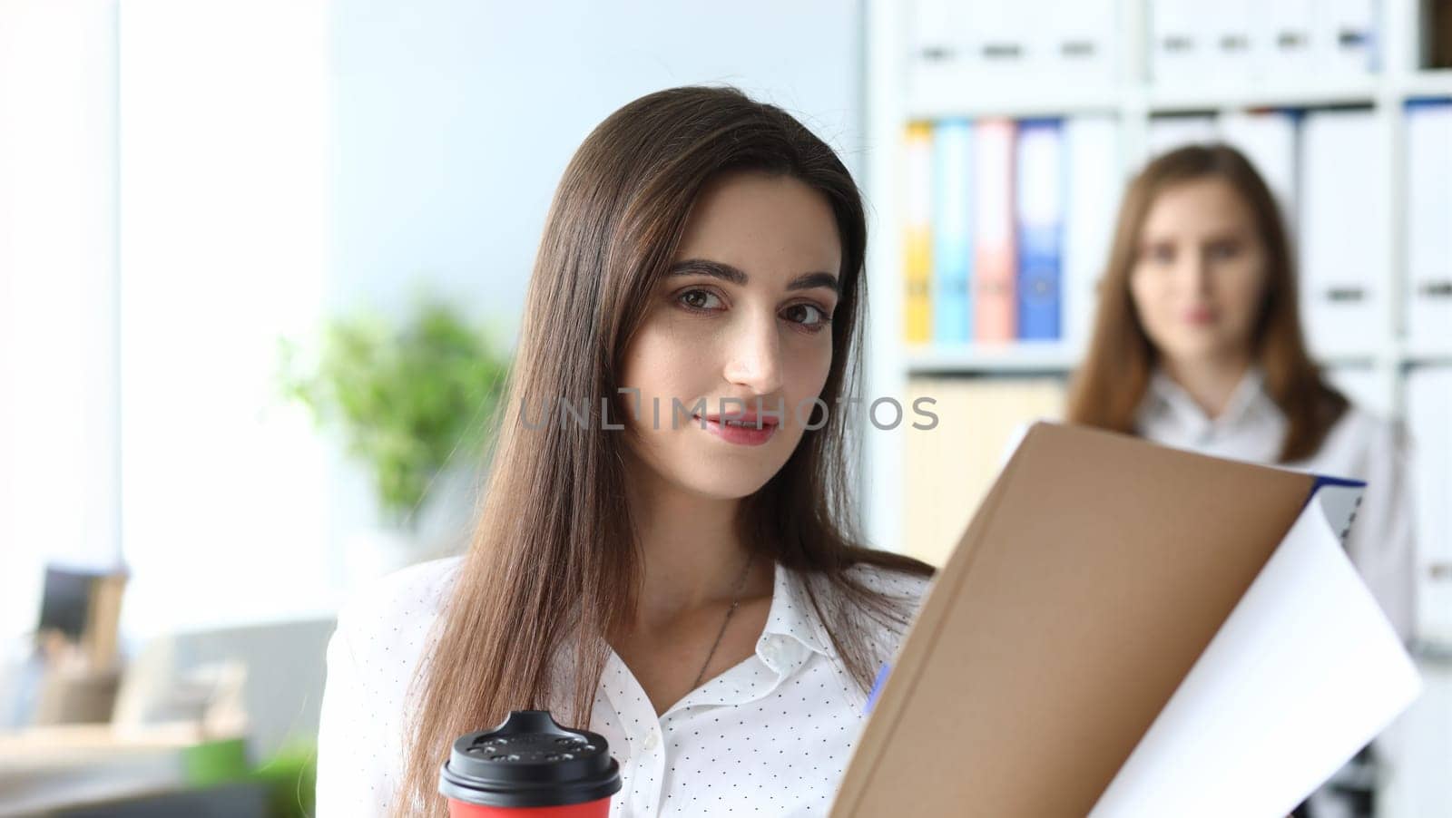 Portrait of joyful businesswoman holding red paper coffee cup and important tablet with significant documents with smart friendly hardworking female colleague. Accounting office concept