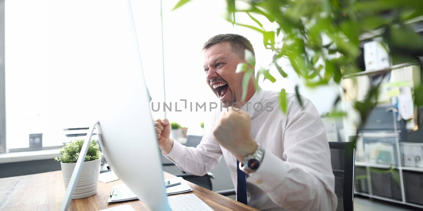 Portrait of cheerful man looking at computer monitor with gladness and making successful gesture. Businessman finishing project and wins big. Blurred background