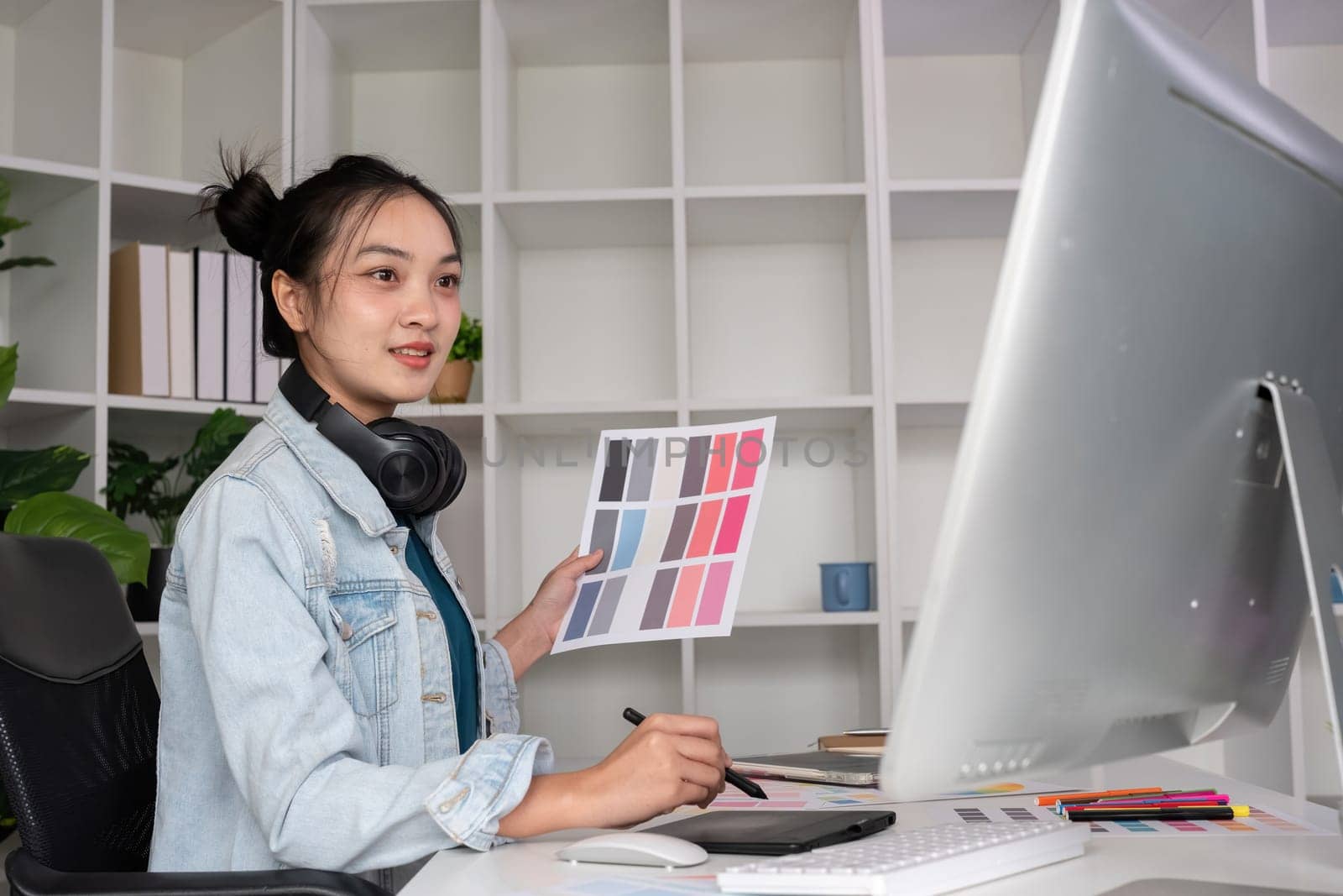 Female designer wearing headphones listening to music while doing graphic design work Choose colors for working with the computer at the table. by wichayada