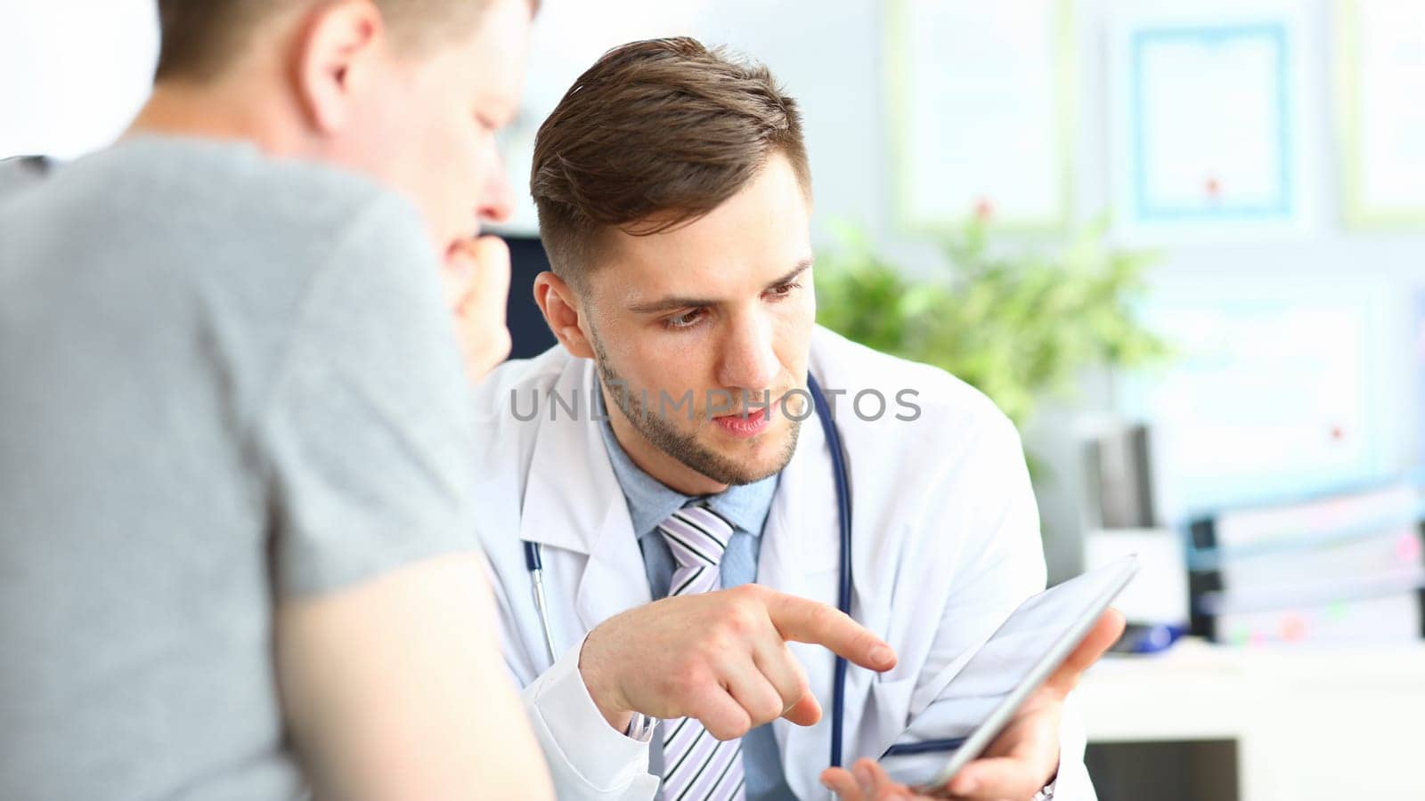 Portrait of concentrated doctor pointing to gadget with finger. Patient listening attentively to doc recommendations. Medical treatment concept. Blurred background