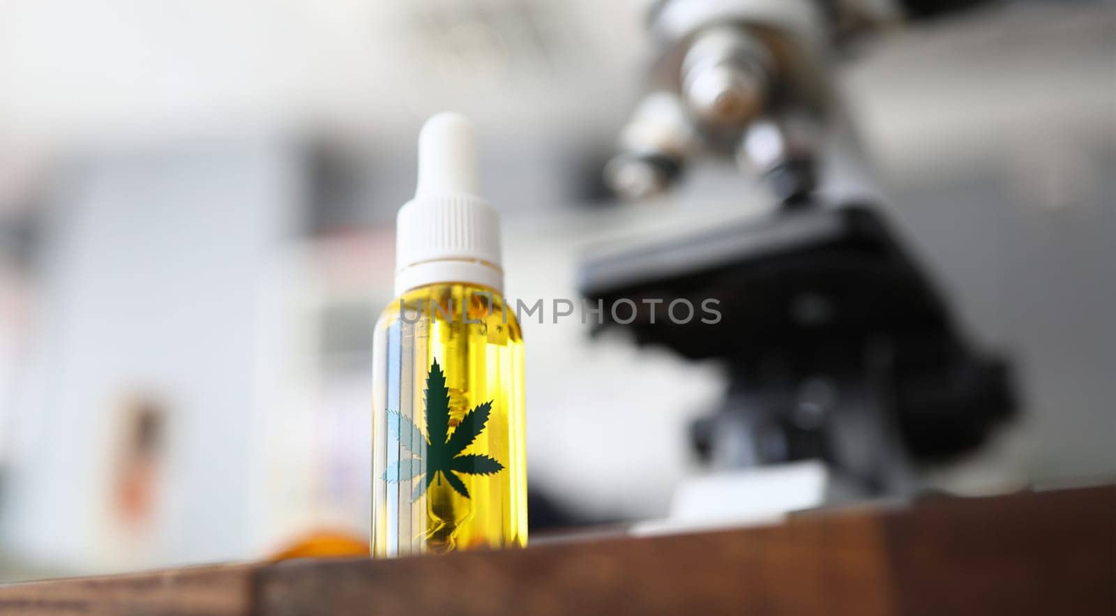 Focus on glass flask with cannabinoid oil and green ganja symbol on it. Laboratory interior with microscope on blurred background. Medicine and medical marijuana concept