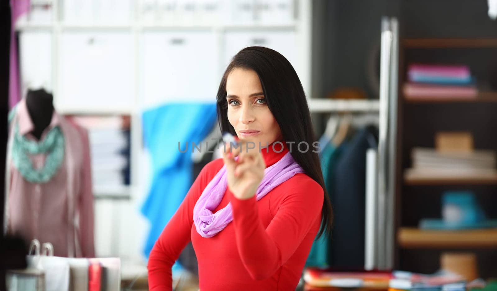 Portrait of beautiful female tailor standing in modern workspace. Smart fashion professional wearing red stylish outfit and pointing at camera with pen. Fashioner workshop concept