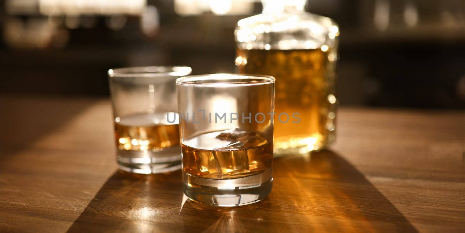 Focus on two glassful and bottle with whiskey or bourbon on wooden table. Luxury drink with ice-cubes on desk. Celebration concept. Blurred background