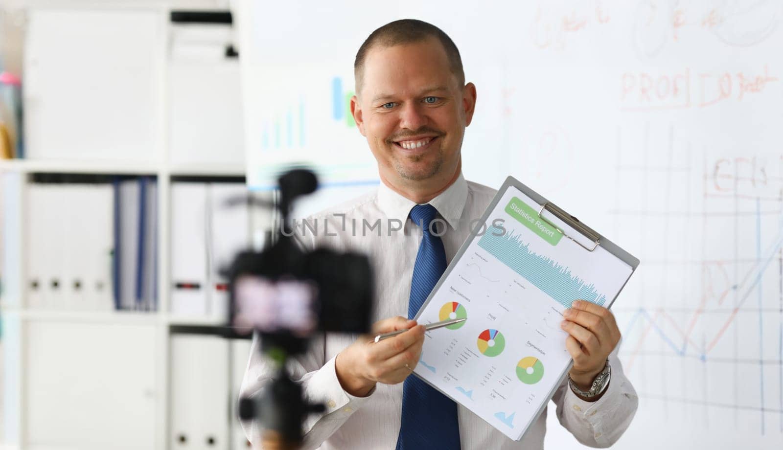 Portrait of smiling blogger in classic suit and tie showing statistics data with graphs charts diagram. Promo videoblog or photo session in office camcorder to tripod. Blurred background