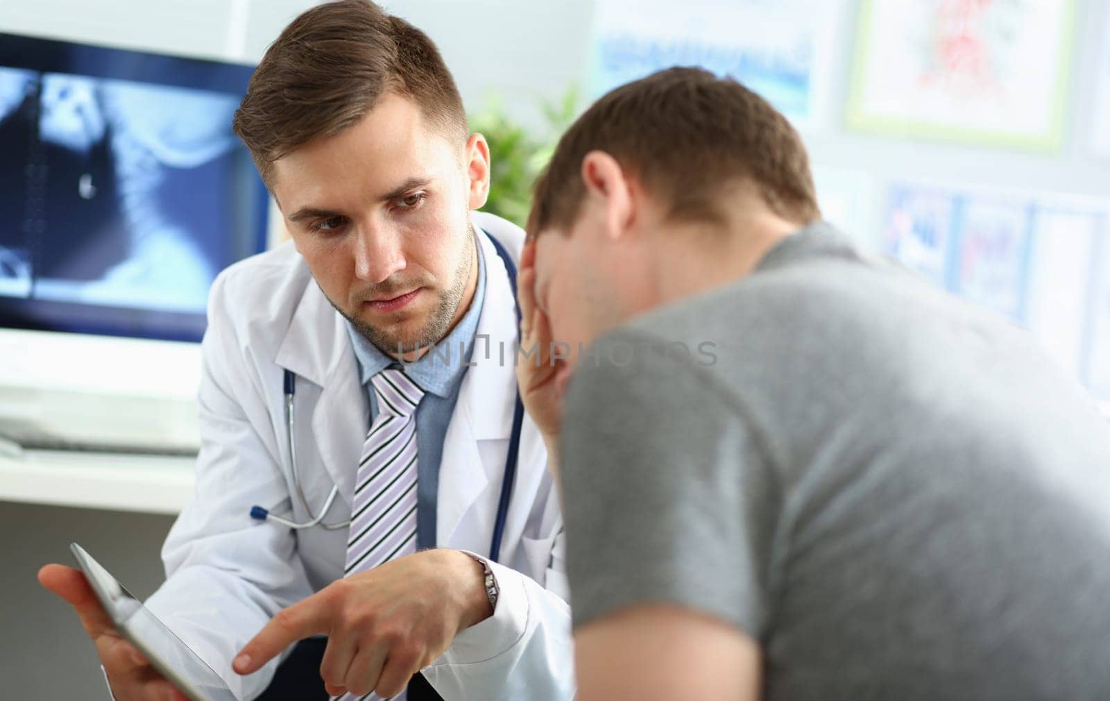Portrait of doctor talking with patient. Physician pointing to something on tablet and seriously looking at sad visitor. Upset male sitting with head down. Medicine concept. Blurred background