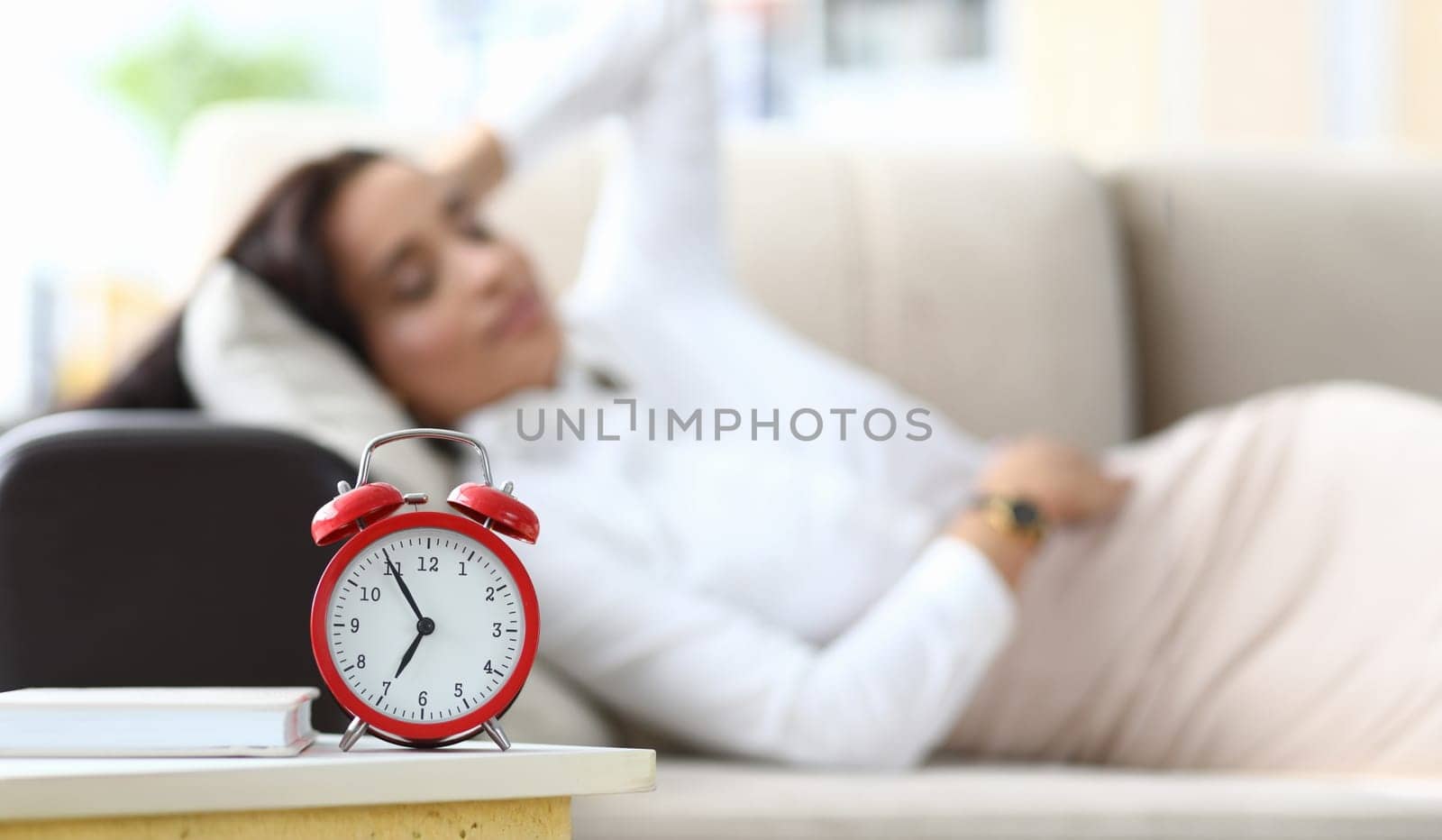 Focus on red alarm clock put near comfortable sofa of tired businesswoman lying on cozy divan and redusing high level of stress with sleeping. Chilling on coach concept