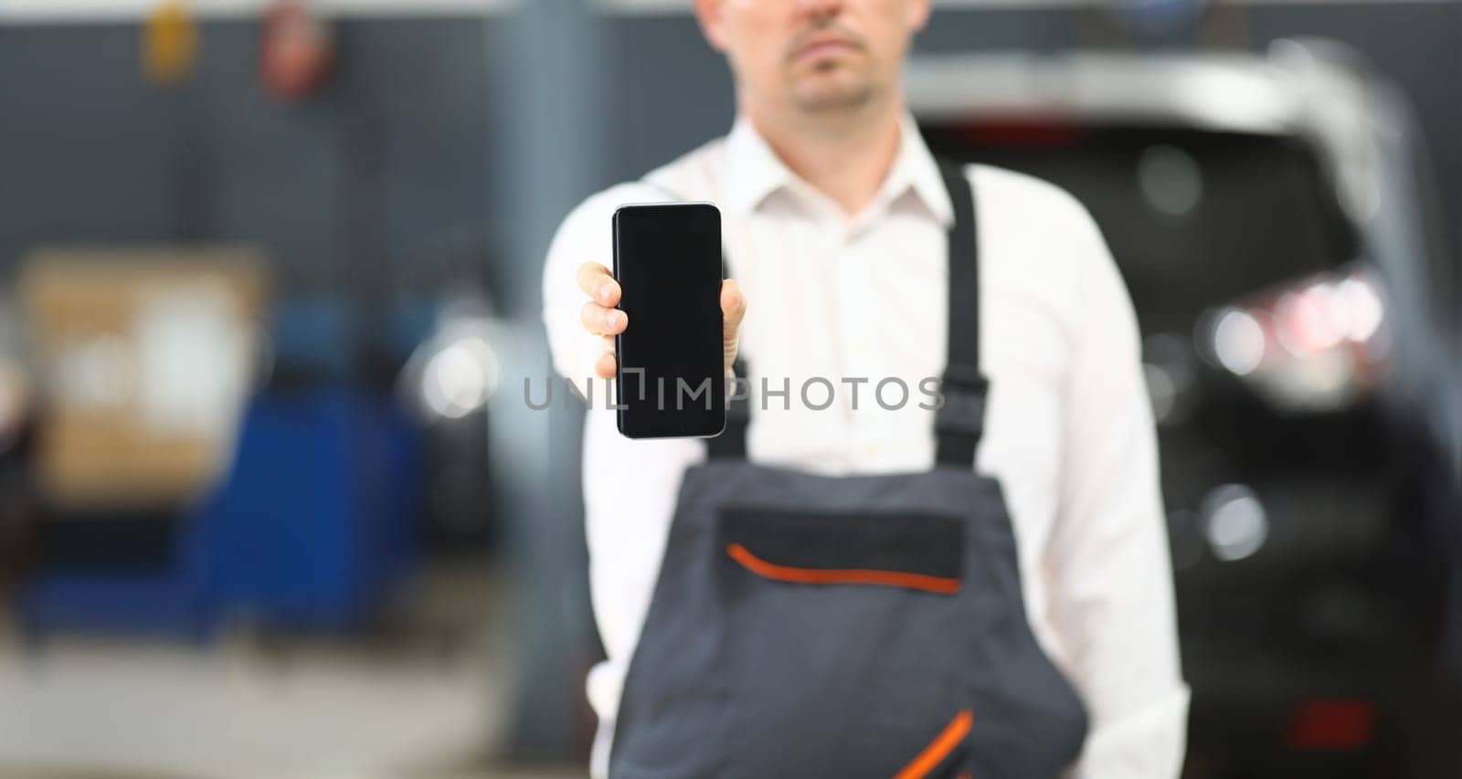 Focus on car repairing engineer standing in modern garage and demonstrating high-tech smartphone on camera. Empty copy space on screen. Blurred background