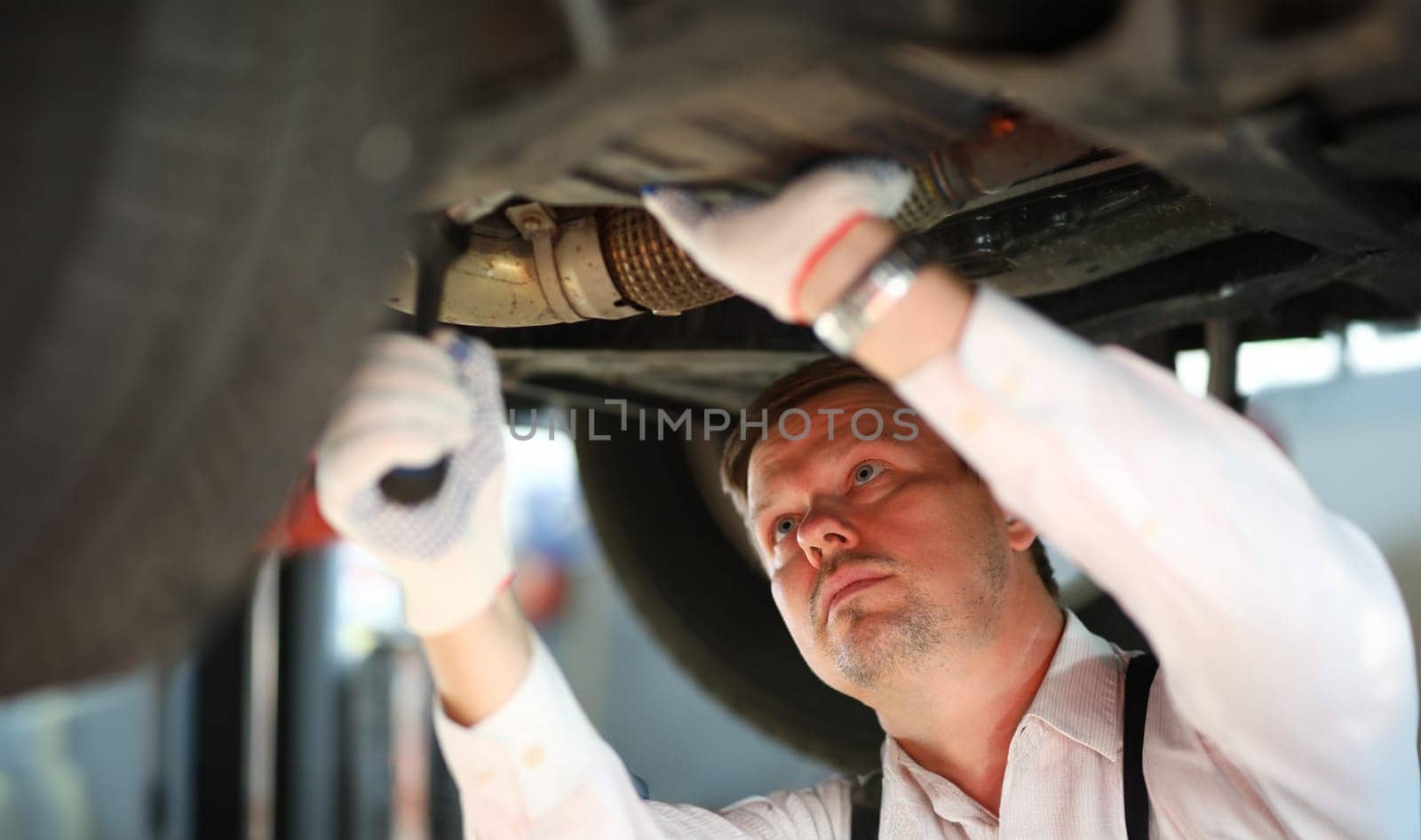 Portrait of hardworking man standing in garage and looking at underneath pipes of modern automobile in white gloves. Machinery repairman and service station concept