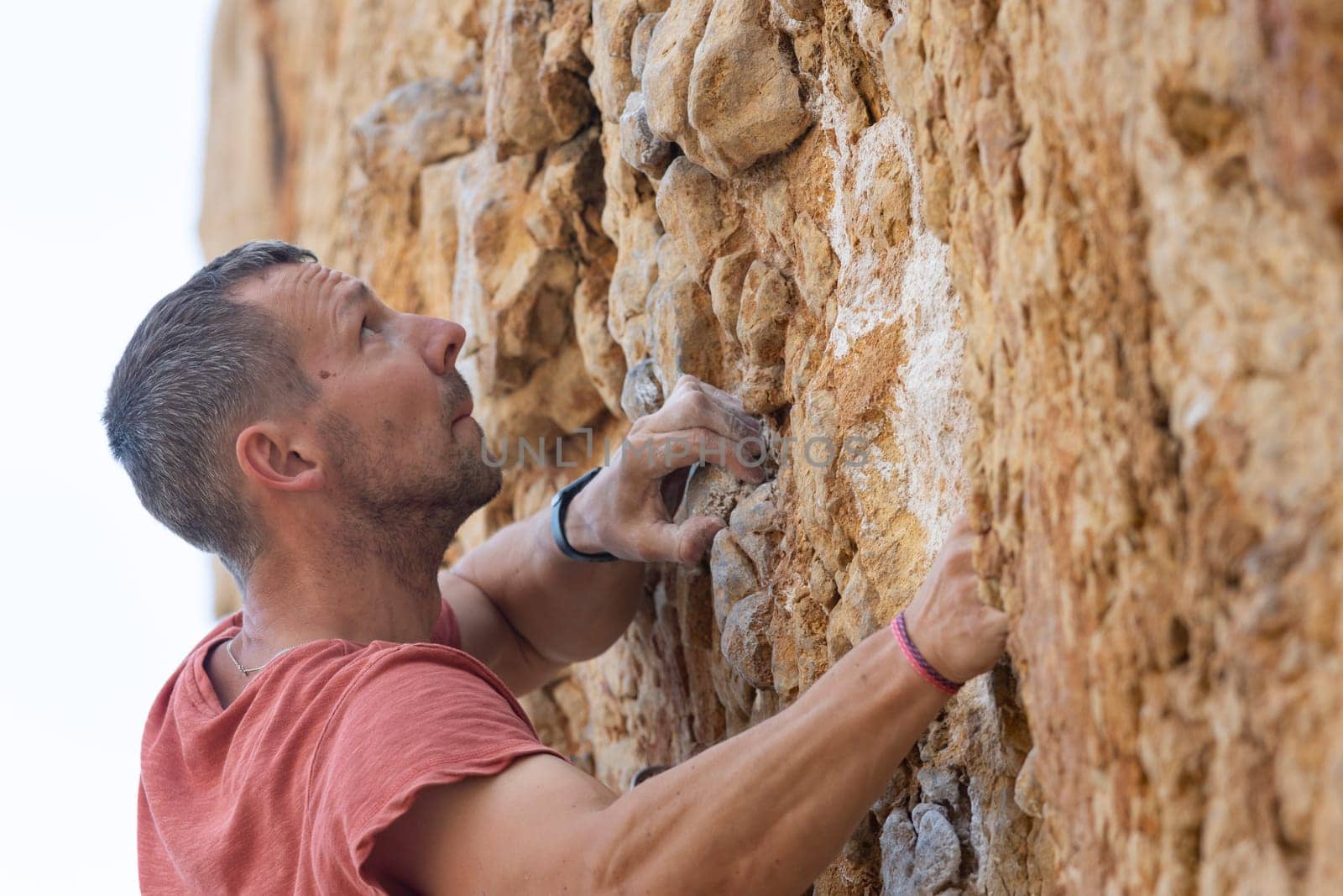 A man in a red shirt is climbing a rock wall. He is wearing a black wristband and a red bandana