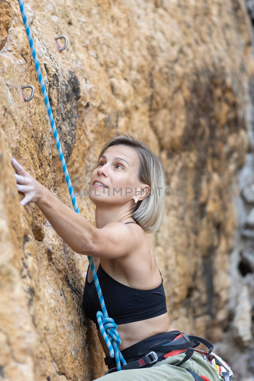 A woman is climbing a rock wall with a blue rope. She is wearing a black tank top and is looking up at the wall