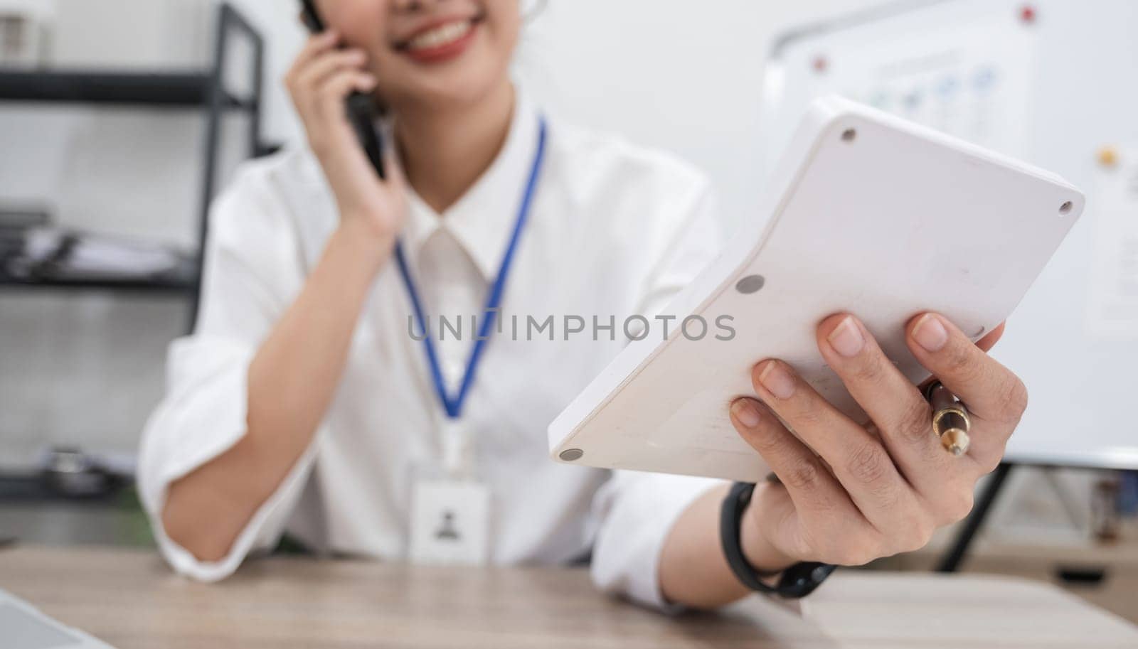 Asian female accountant is talking on the phone in the office with working documents regarding calculations on the office table. by wichayada