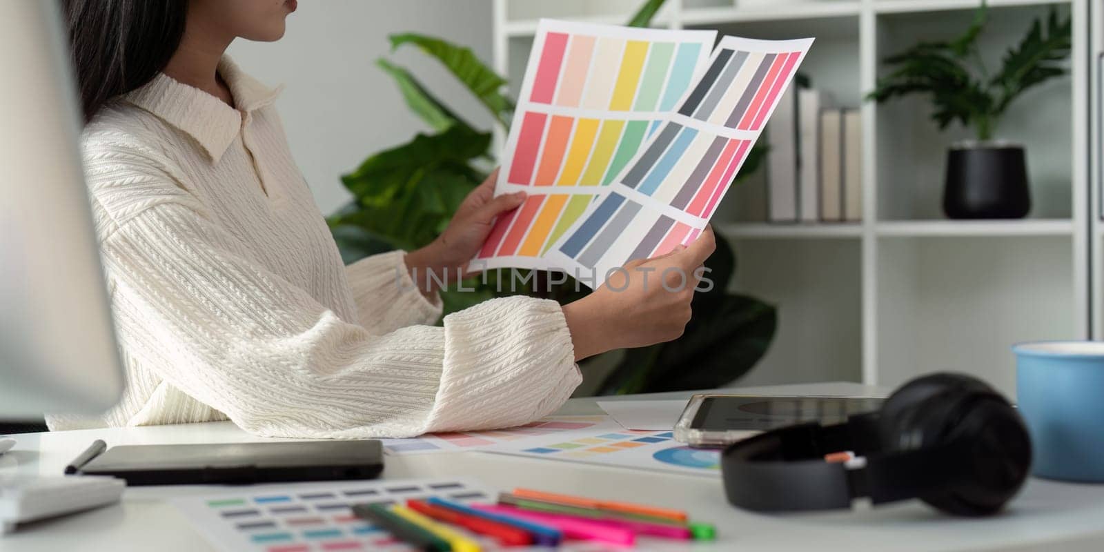 Asian woman freelance graphic designer working with color swatch samples and computer at desk in home office, young lady choosing color gamma for new design project.