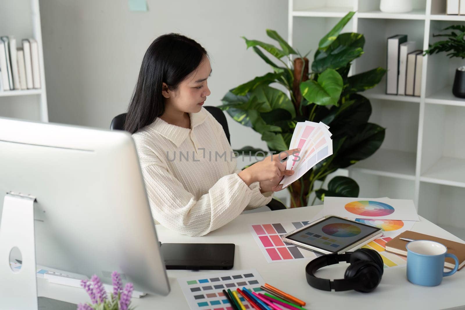 Asian woman freelance graphic designer working with color swatch samples and computer at desk in home office, young lady choosing color gamma for new design project by nateemee