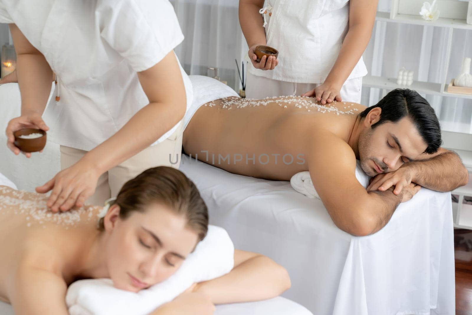Blissful couple customer having exfoliation treatment in luxury spa salon with warmth candle light ambient. Salt scrub beauty treatment in health spa body scrub. Quiescent