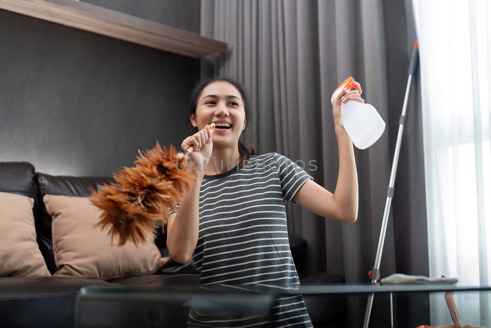 House cleaning with fun. Happy young asian housewife singing song during cleanup, using feather duster as microphone, enjoying domestic work. Young woman dancing and cleaning in living room.