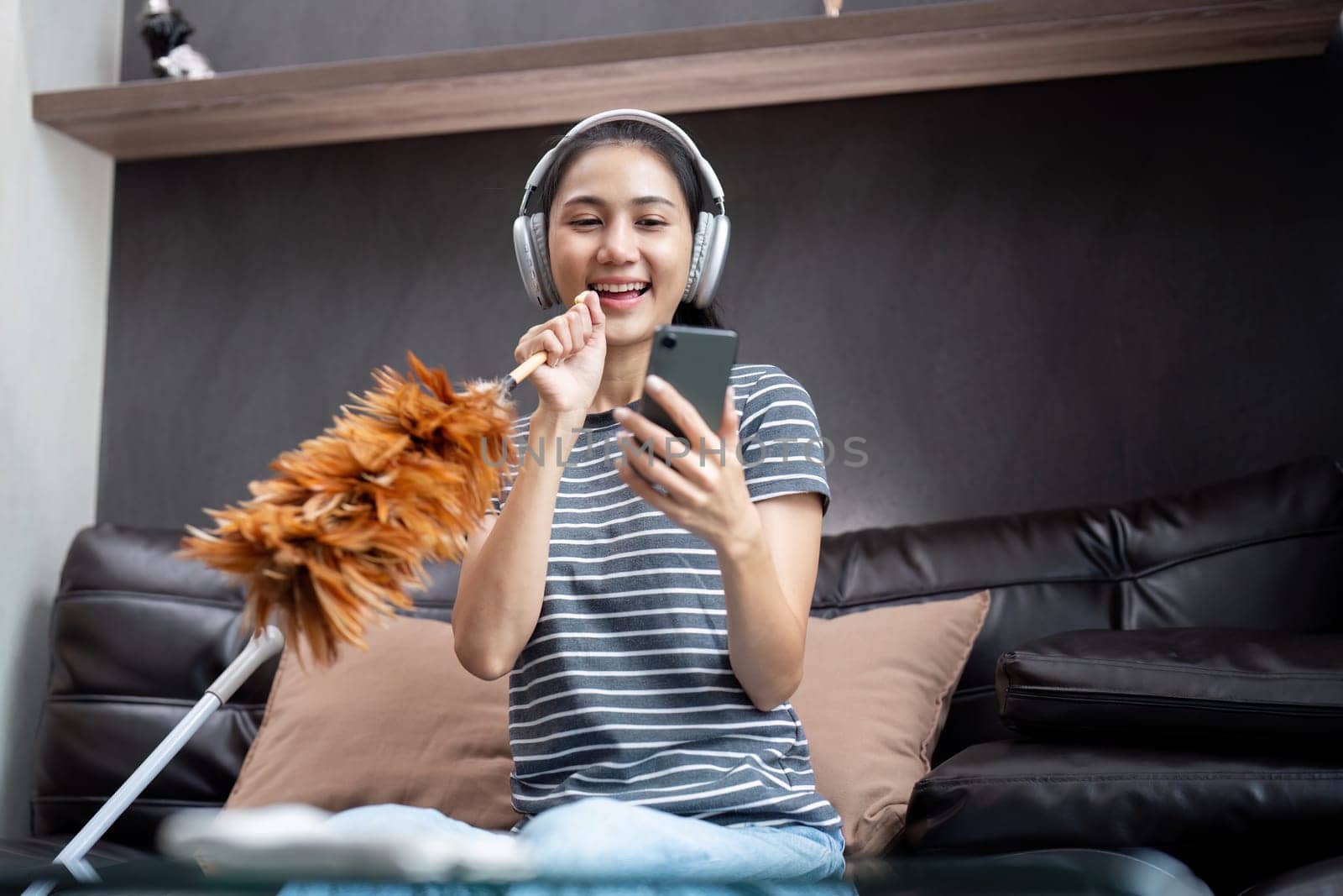 House cleaning with fun. Happy young asian housewife singing song during cleanup, using feather duster as microphone, enjoying domestic work. Young woman dancing and cleaning in living room.