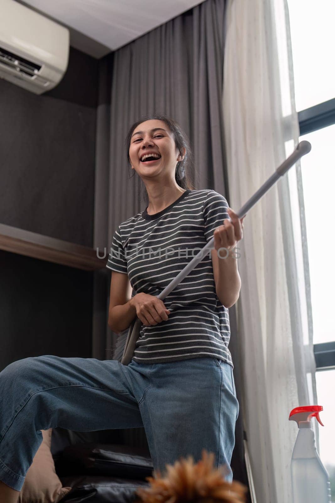 House cleaning with fun. Happy young asian housewife singing song during cleanup, using mop as guitar, enjoying domestic work. Young woman dancing and cleaning in living room by nateemee