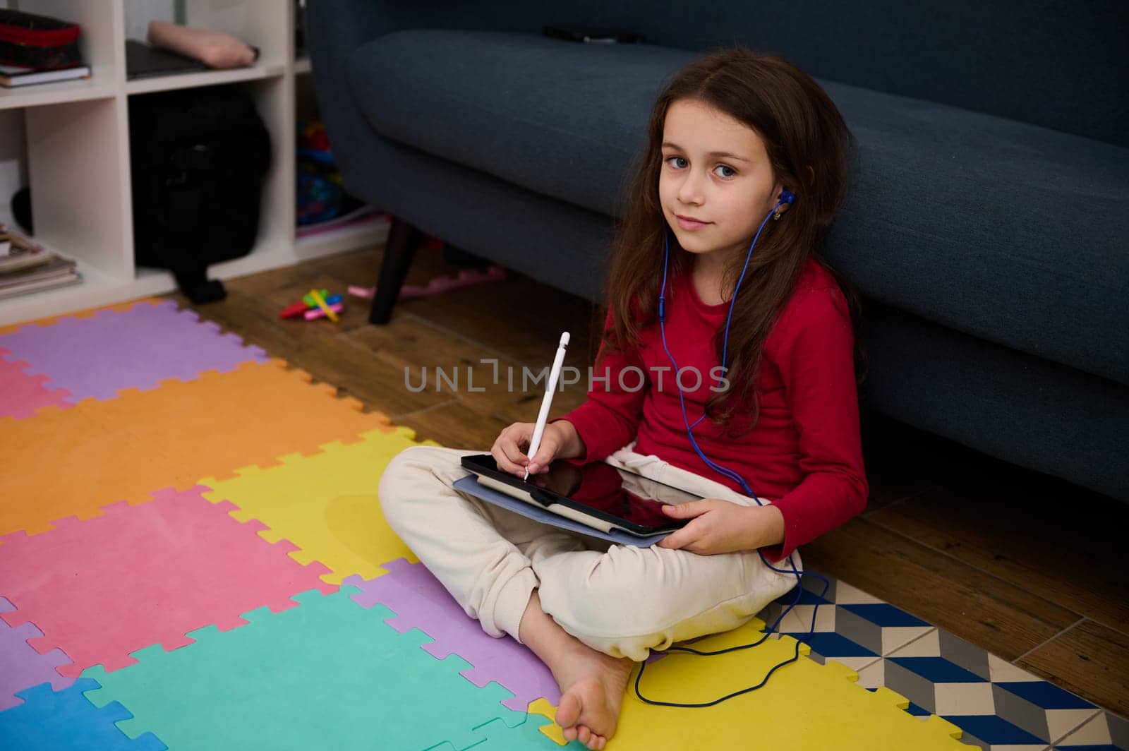 Delighted little child girl in casual clothes and headphones, smiling for camera and listening to music while sitting crossed legs on multicolored puzzle carpet and using a digital tablet on
